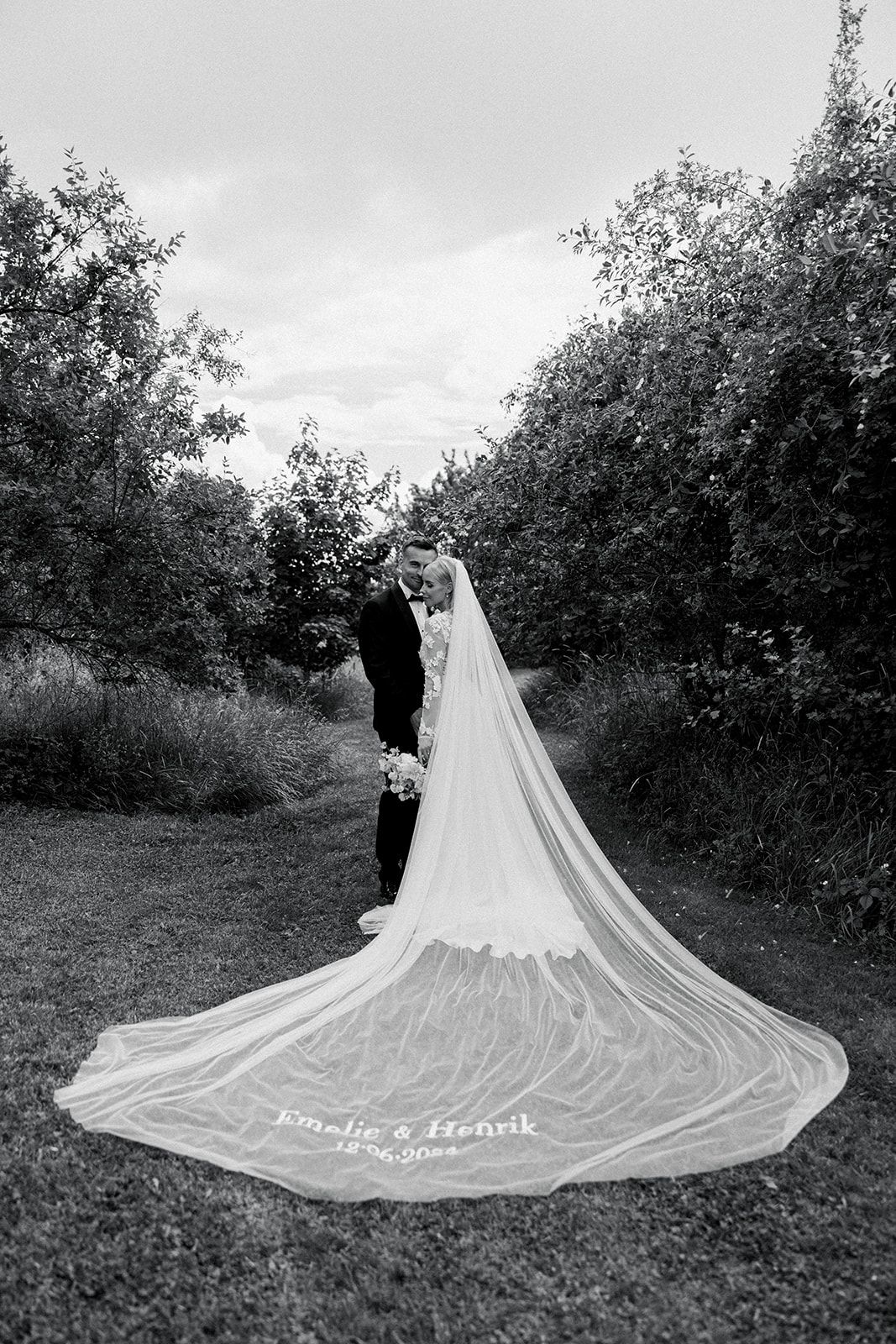 A couple in wedding attire poses in a garden setting with a long flowing cathedral veil trailing behind them.