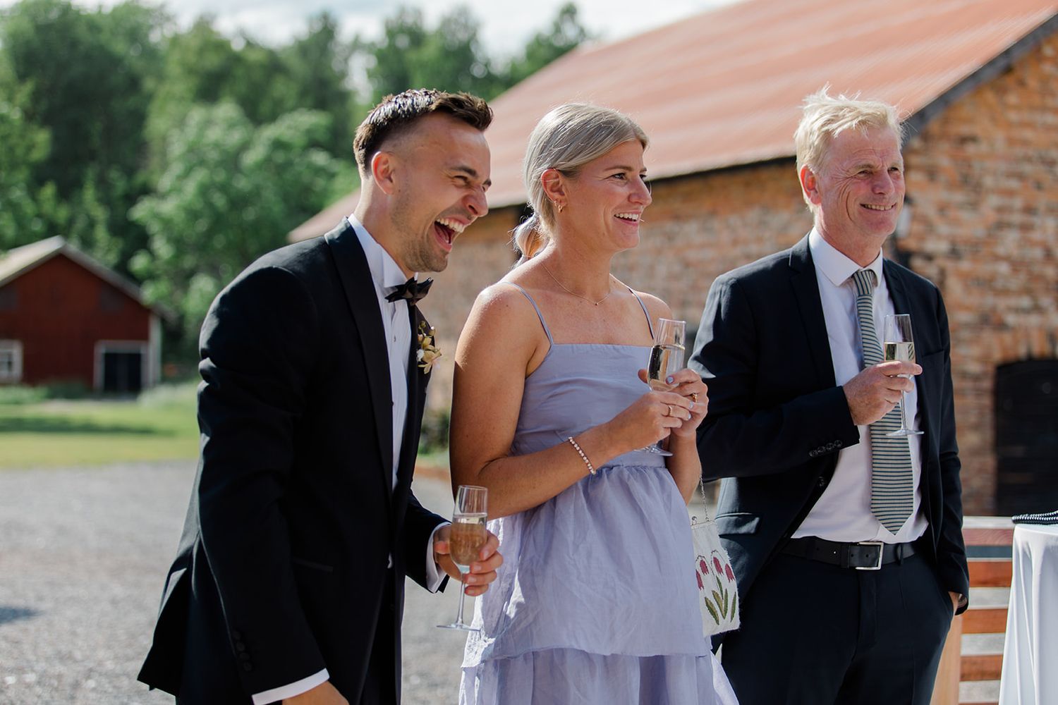 Three guests in formal attire share laughs and drinks at an outdoor wedding reception near a rustic barn.