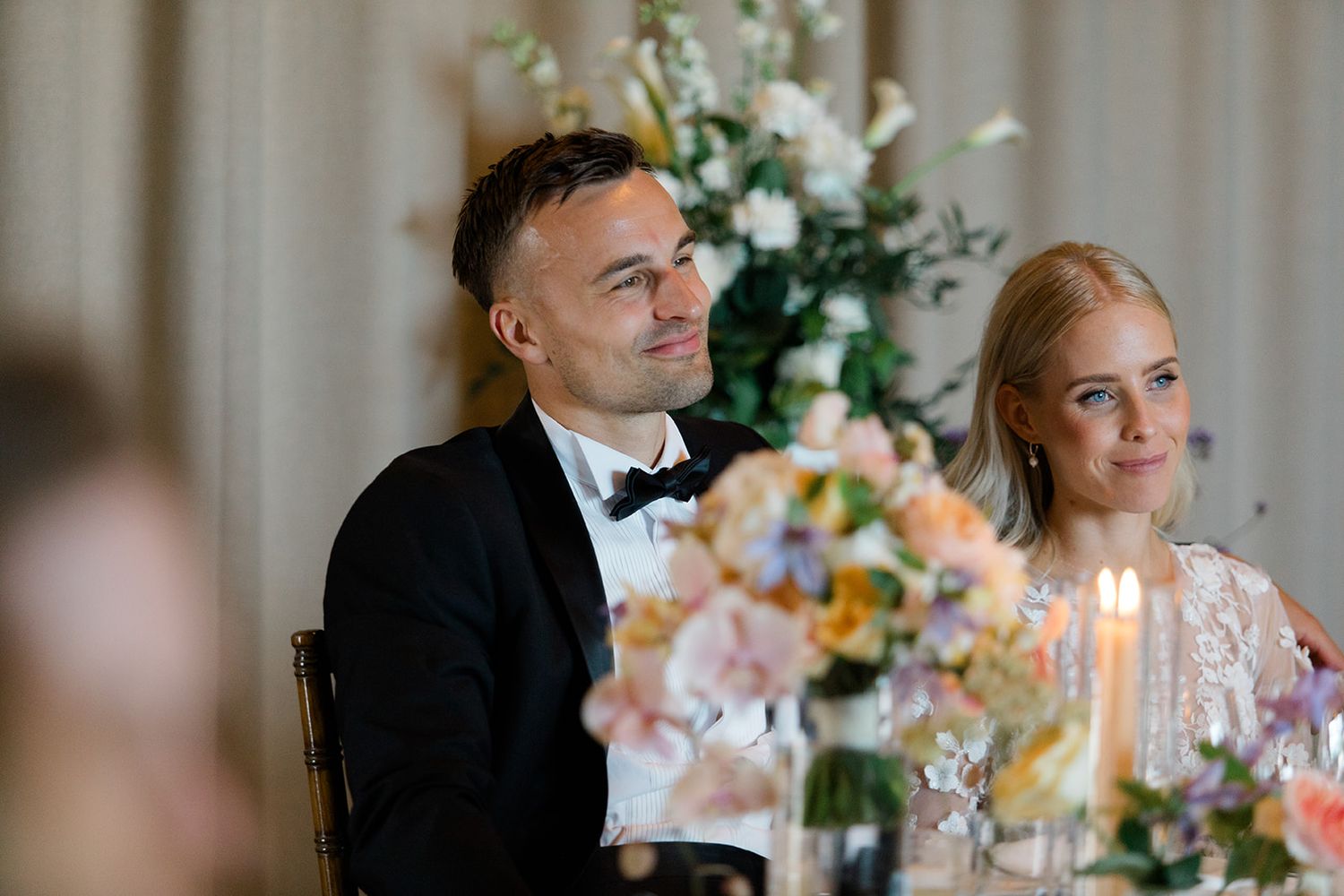 A couple sits at an elegant reception table with floral arrangements and candlelight during a formal dinner event.