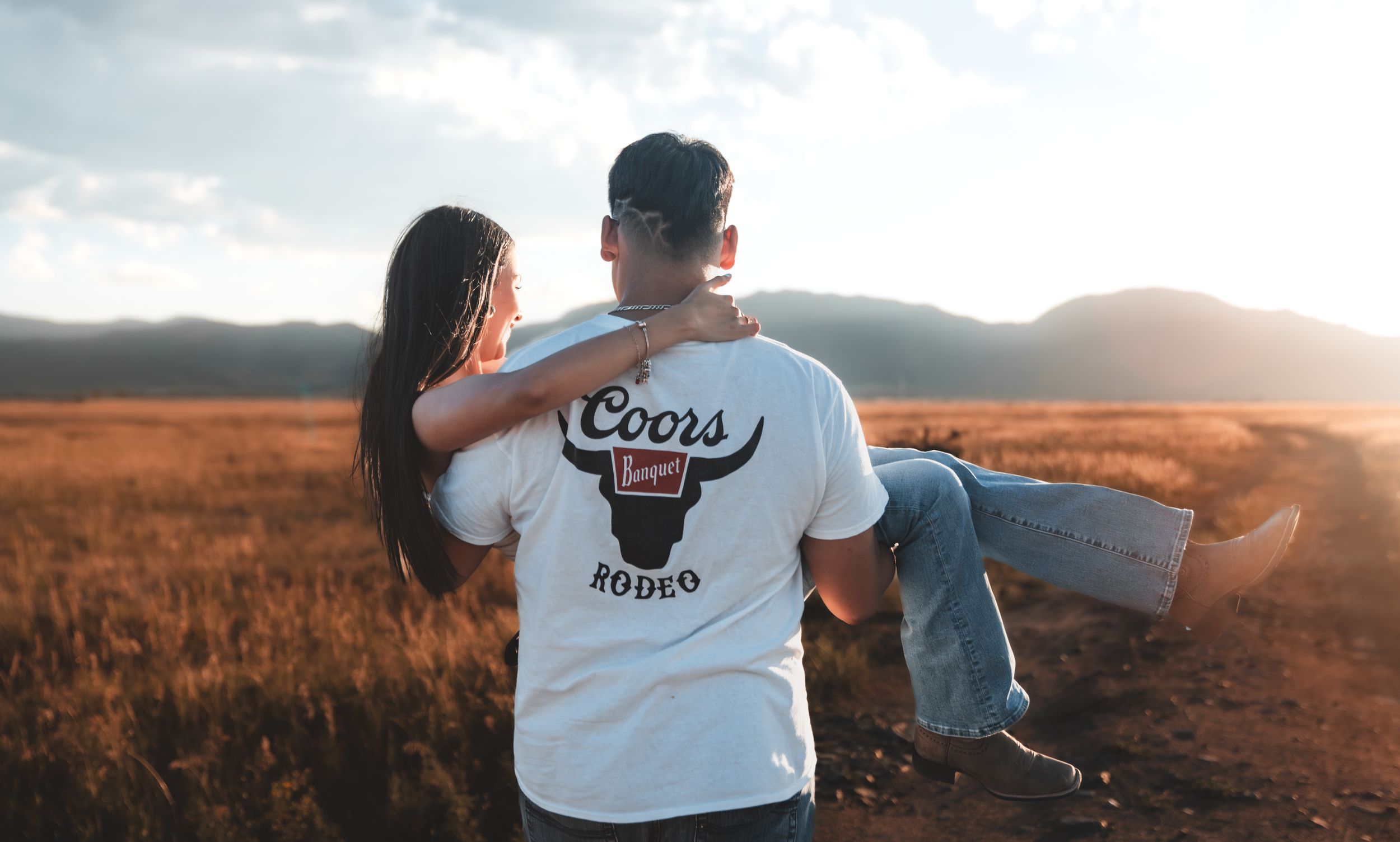 A couple embraces while standing in a golden field during sunset with mountains in the background.