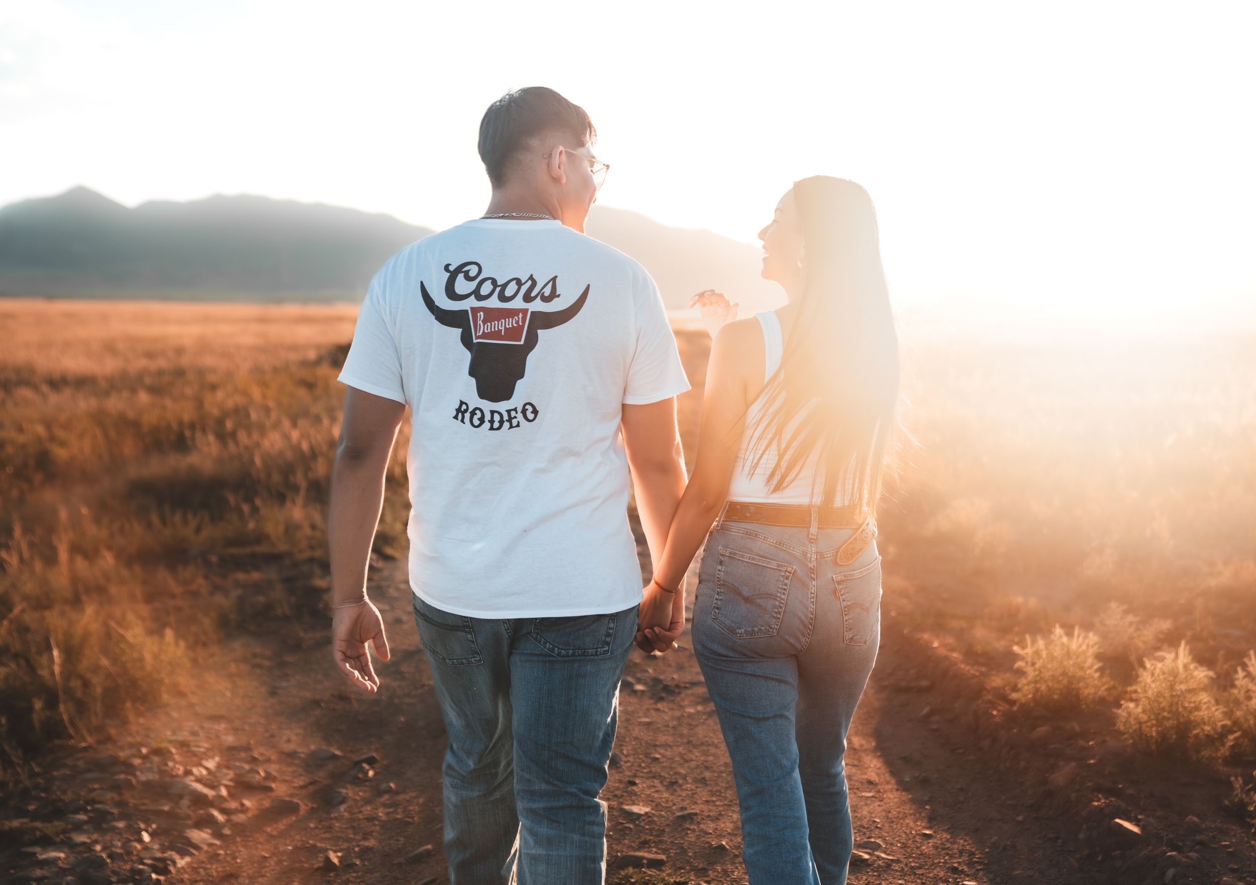 Couple holding hands while walking through a golden field at sunset with lens flare.