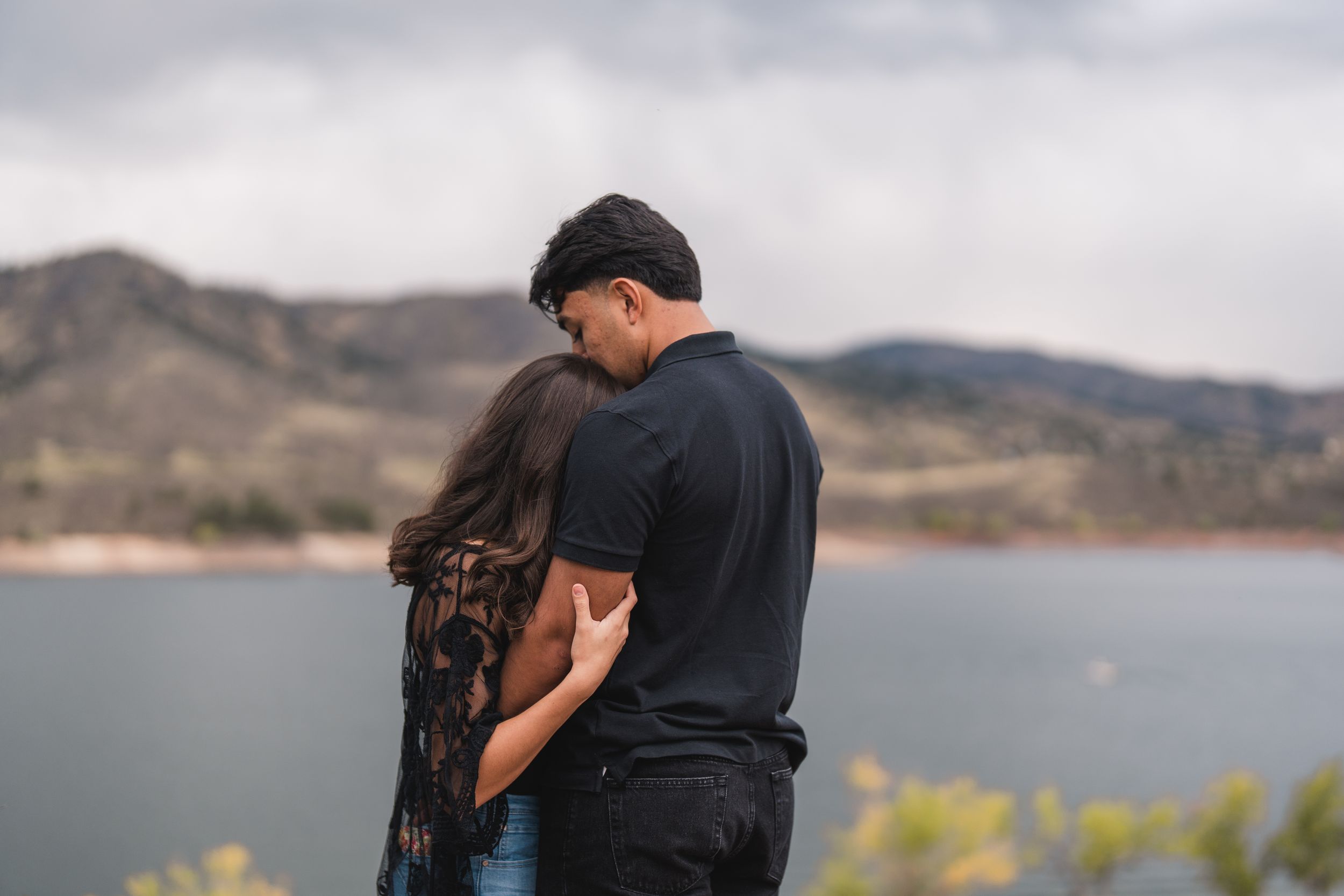 Silhouetted couple embraces while standing on a rocky cliff overlooking a scenic mountain lake at sunset.