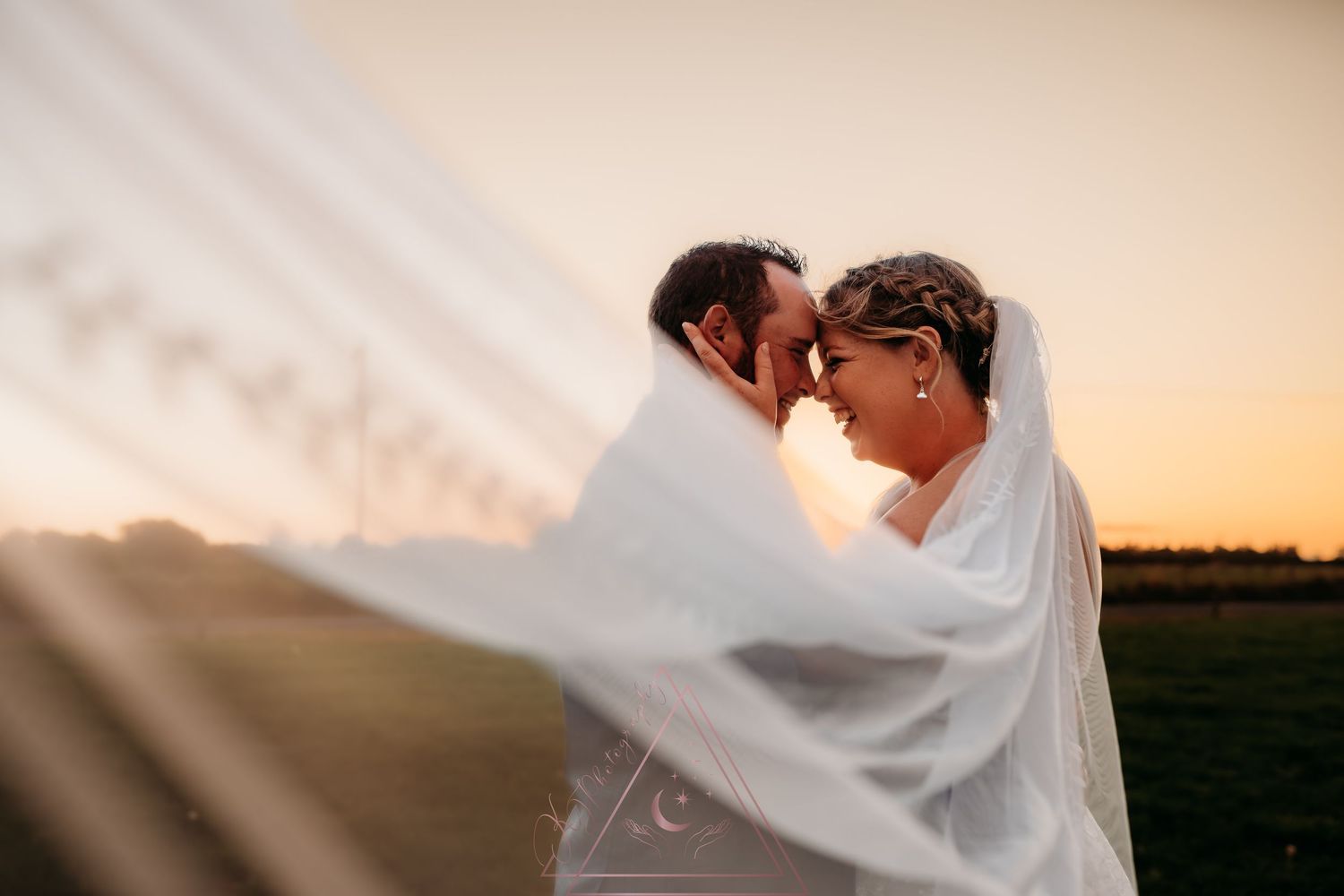 A romantic couple shares a kiss under a flowing white veil at sunset in an outdoor field setting.