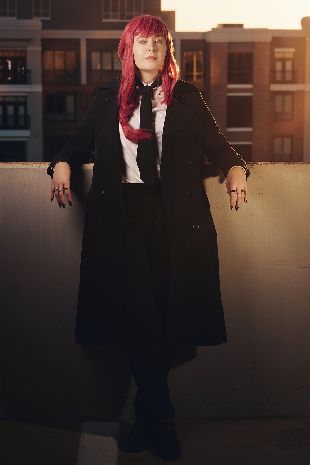A figure in a black coat and bright red hair stands in a dimly lit urban setting with warm sunset lighting.
