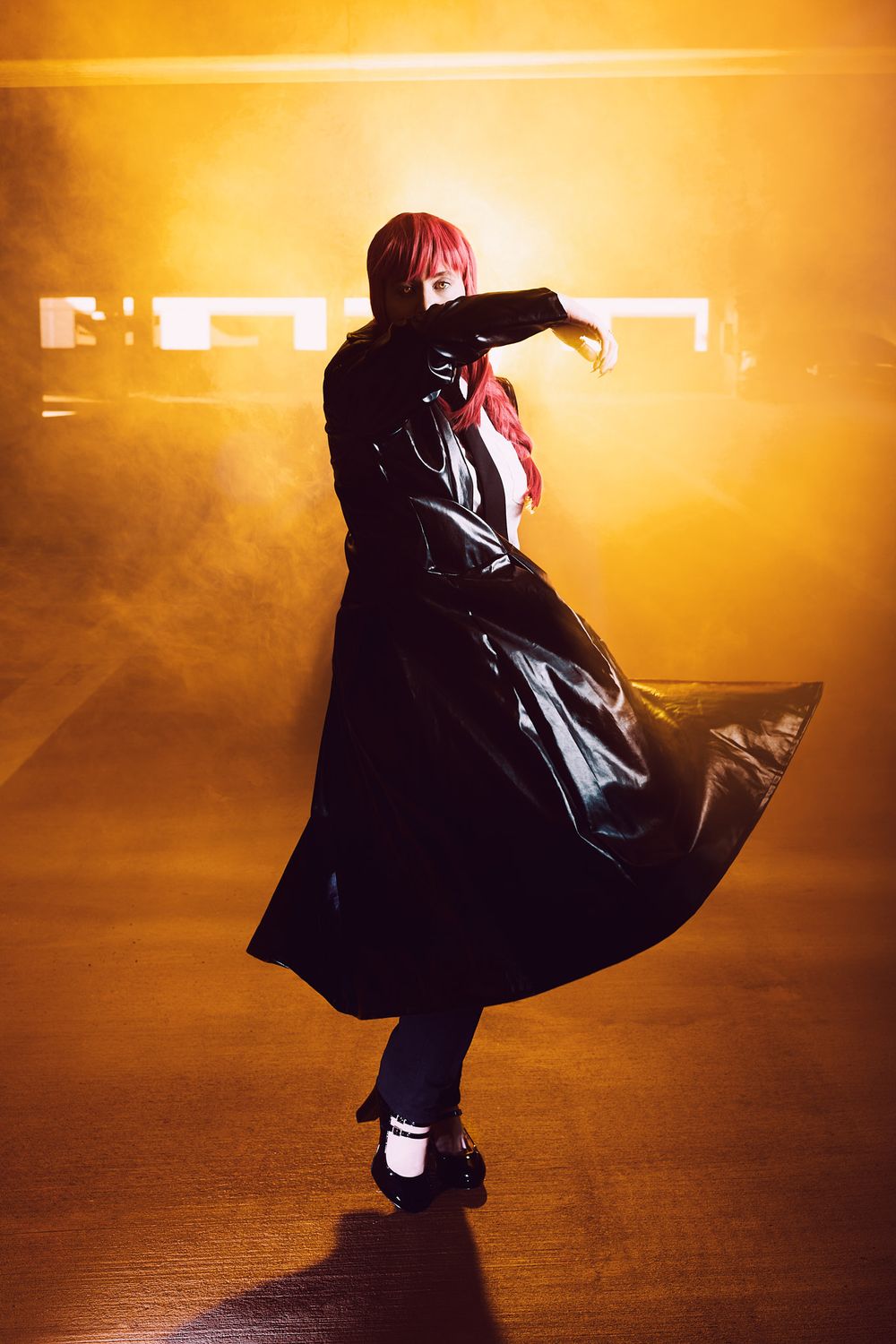 A figure in a flowing black coat and red hair dances dramatically in orange-lit atmospheric fog.