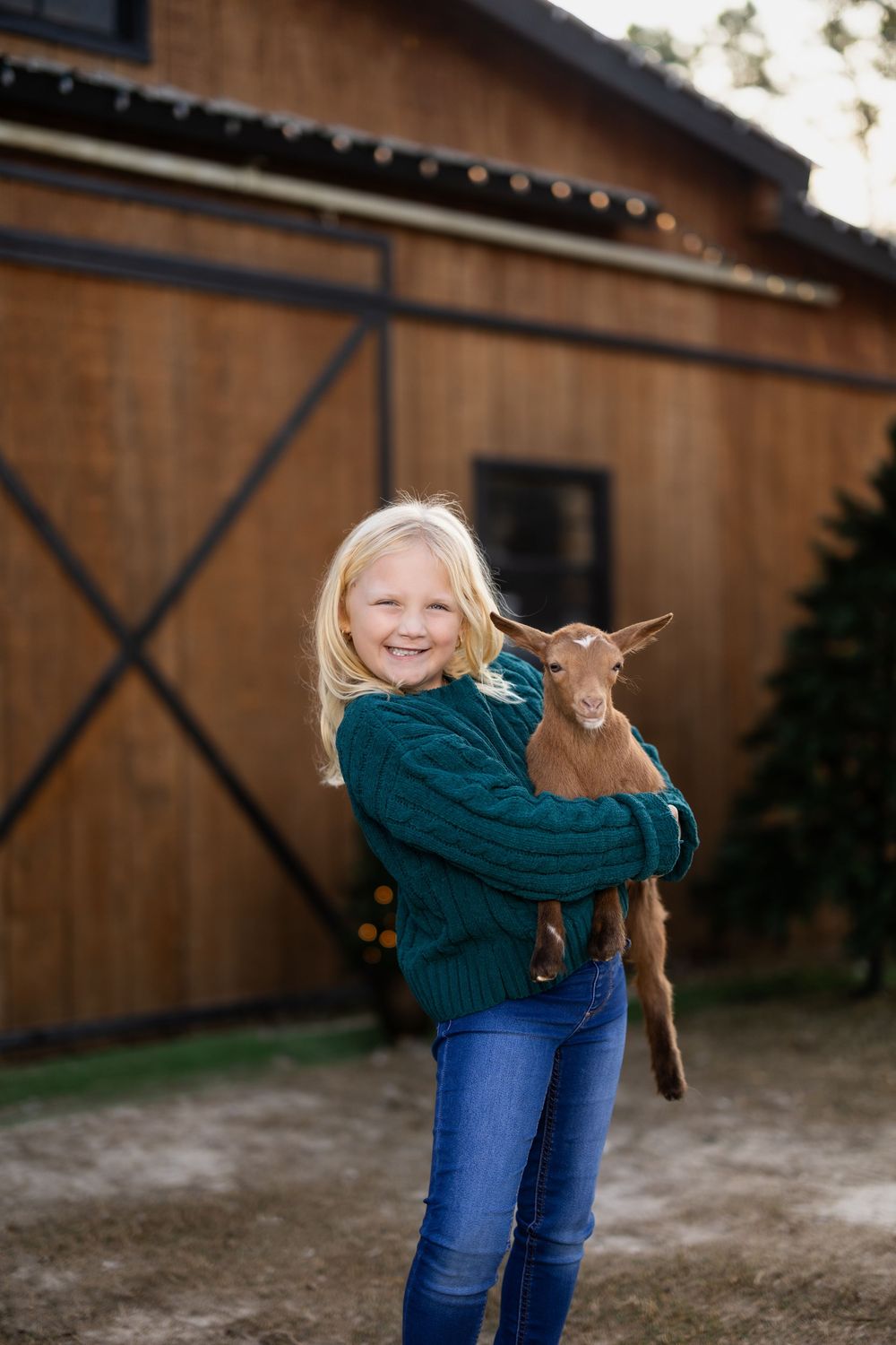 Child in green sweater holds baby goat in front of rustic barn.