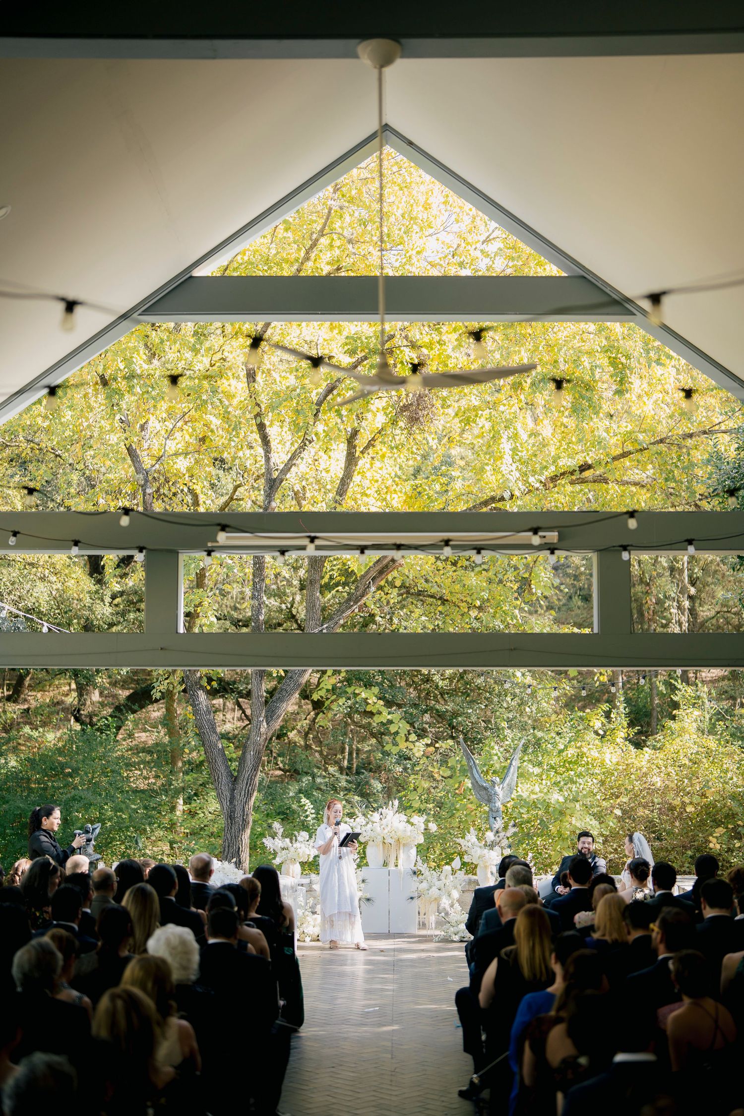 Panoramic view of an outdoor wedding ceremony under a modern pavilion surrounded by autumn trees and natural light.