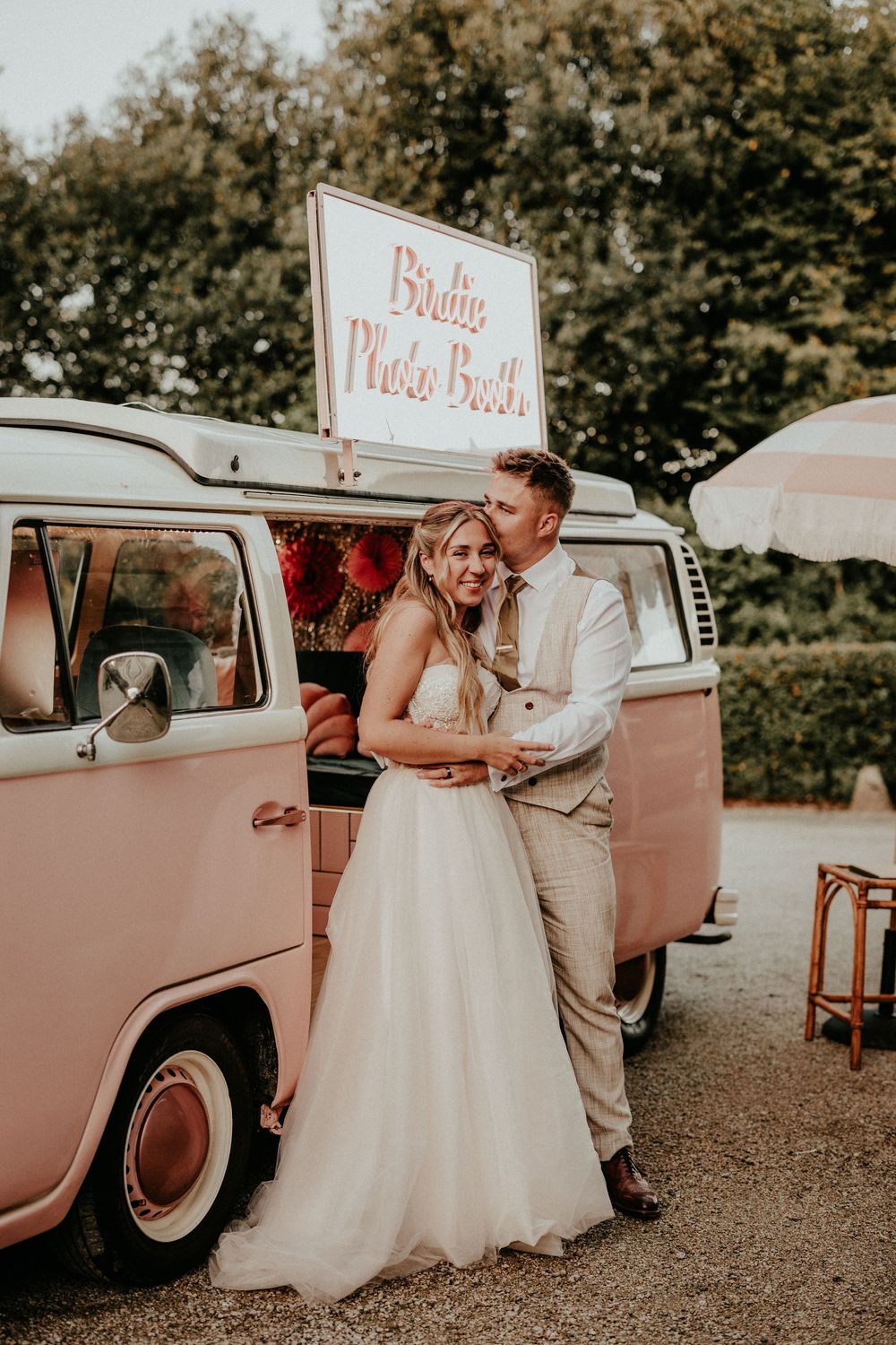 A couple in wedding attire poses beside a vintage pink car with a 'Photo Booth' sign during their outdoor celebration.