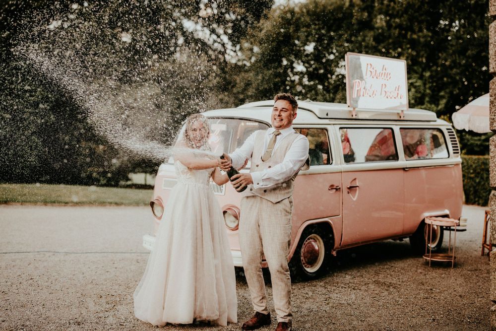 Vintage pink VW bus parked outdoors serves as backdrop for couple popping champagne in wedding attire at sunset. Cornwall wedding photograph