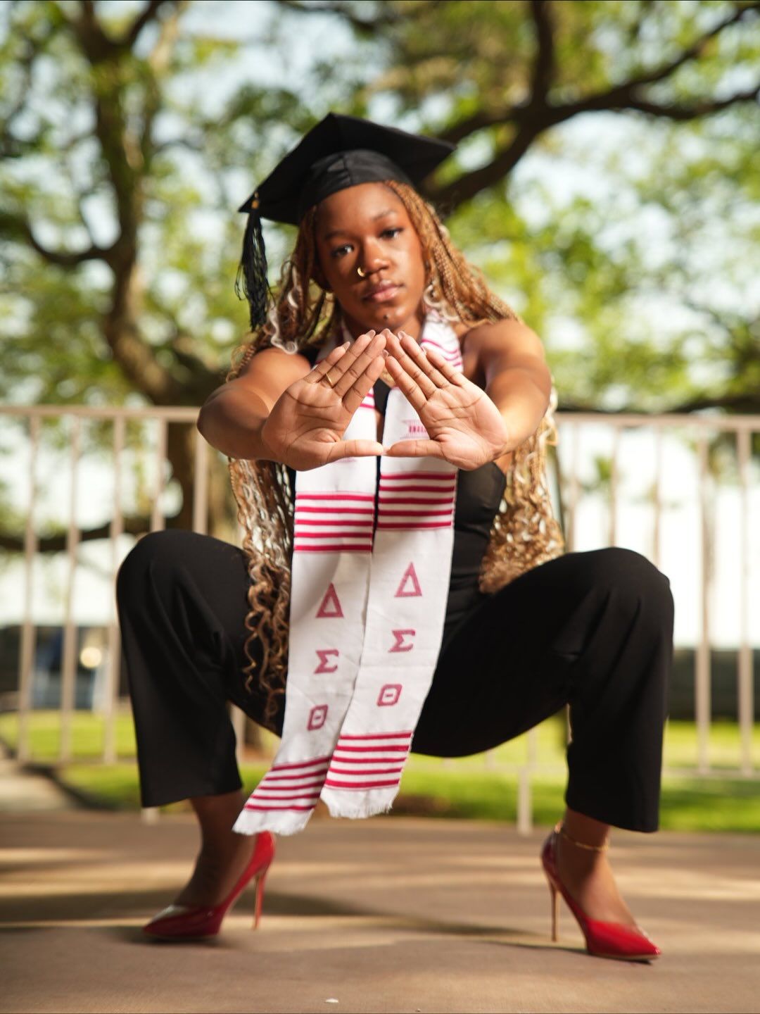 Graduate in a black cap and pink and white Delta Sigma Theta stole crouches outdoors while forming a delta hand sign, wearing red heels.