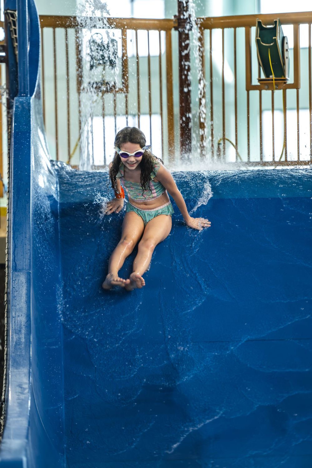 Someone wearing goggles and a swimsuit slides down a blue water slide at a water park with splashing water.