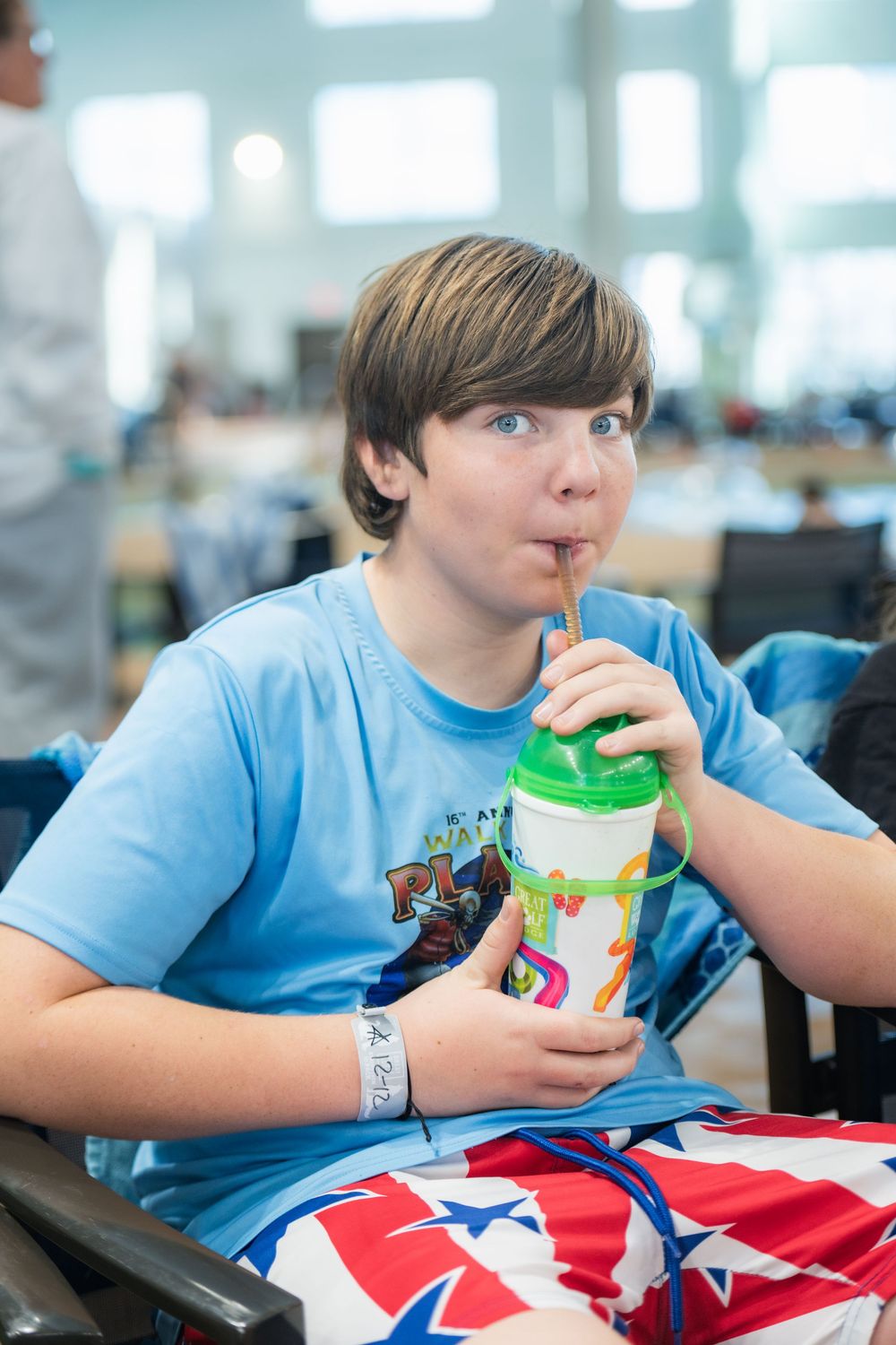 Student in light blue t-shirt drinks from green straw cup during lunch break at school cafeteria.