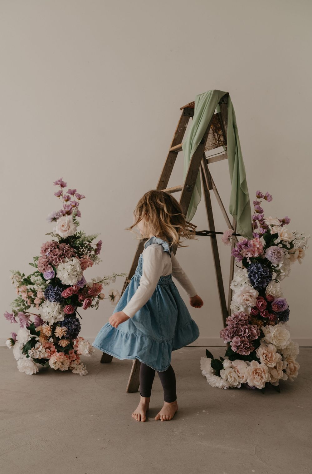 A young girl in a blue dress dances barefoot between floral arrangements flanking a wooden ladder in a studio.