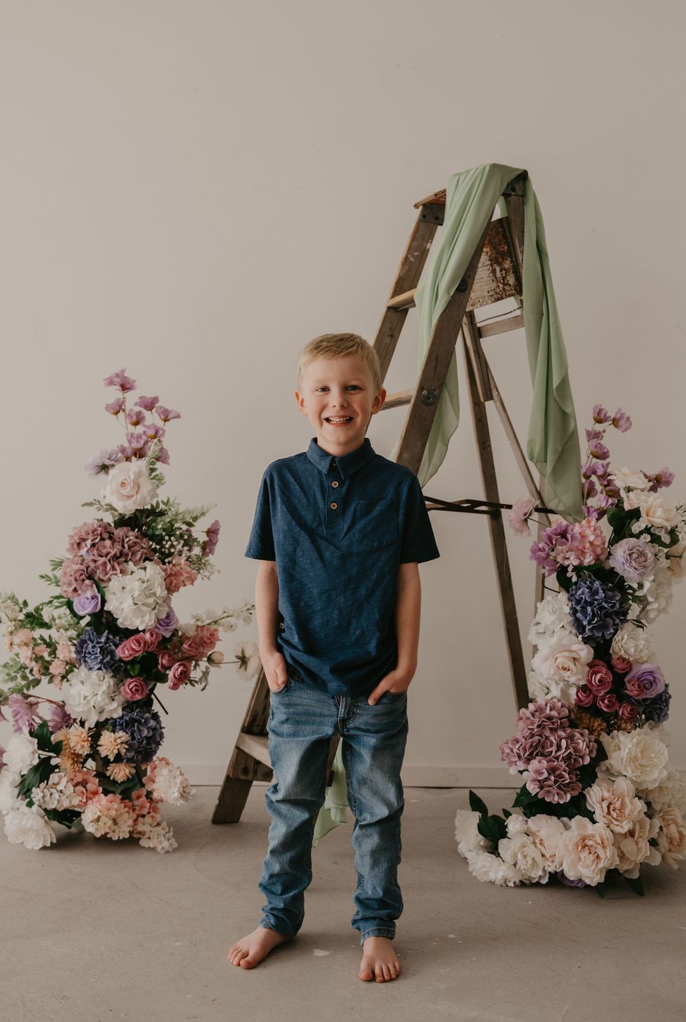 Smiling blonde boy in navy shirt and jeans stands barefoot between floral arrangements beside a rustic wooden ladder.