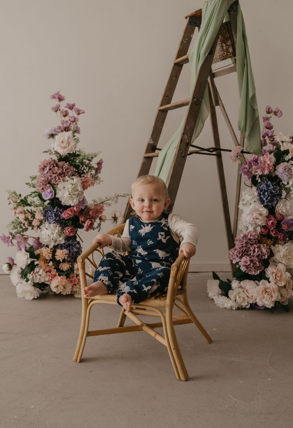 Baby in navy dinosaur romper sits in rattan chair surrounded by lush floral arrangements and wooden ladder backdrop.