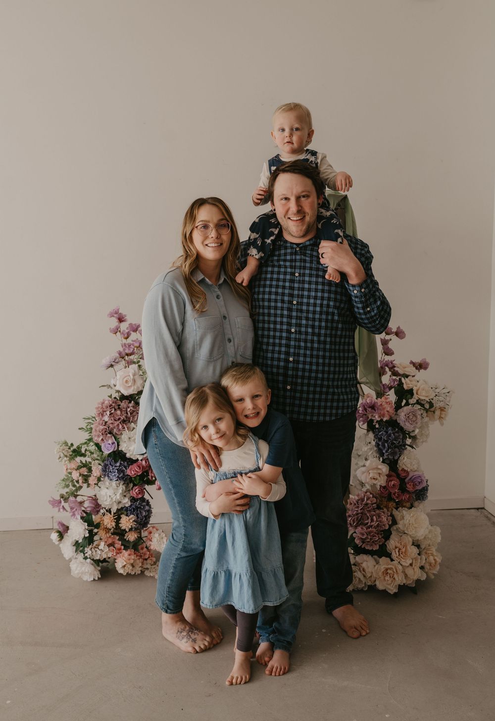 Family of five posing with floral arrangements, parents with three young children in a bright studio setting.
