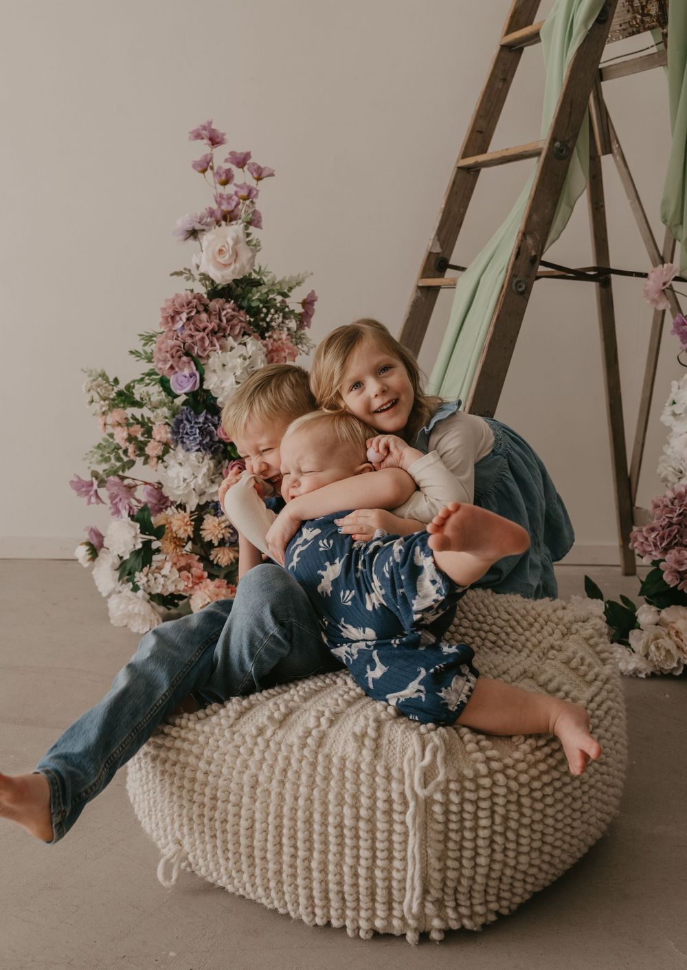 Three young siblings hugging and cuddling together on a woven pouf ottoman surrounded by pastel floral arrangements.