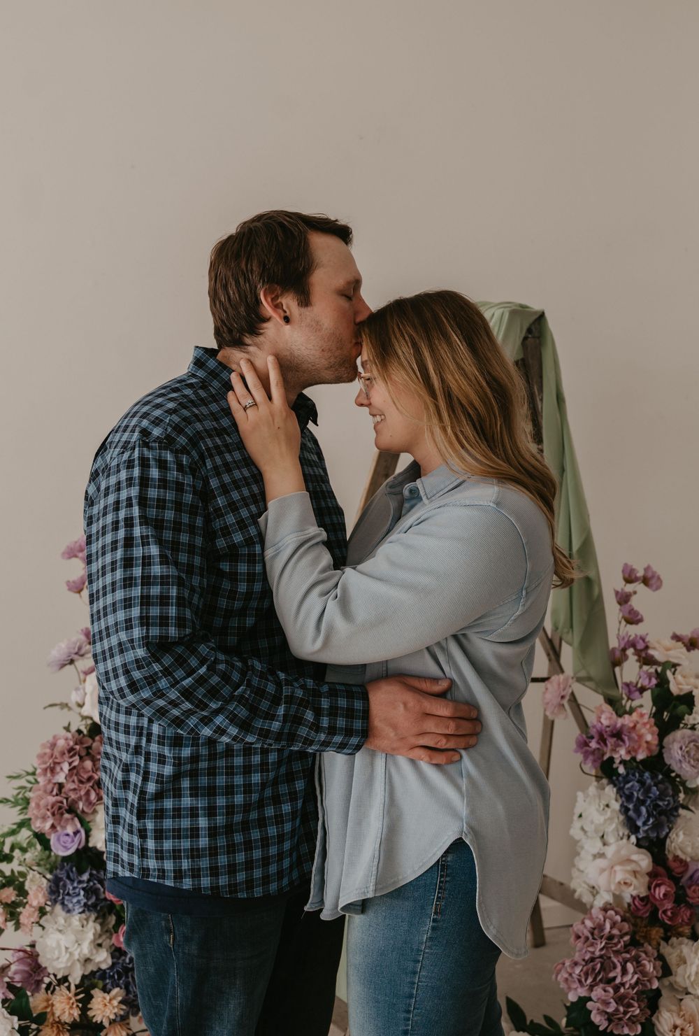 A couple sharing a tender kiss surrounded by purple and pink floral arrangements in a studio setting.
