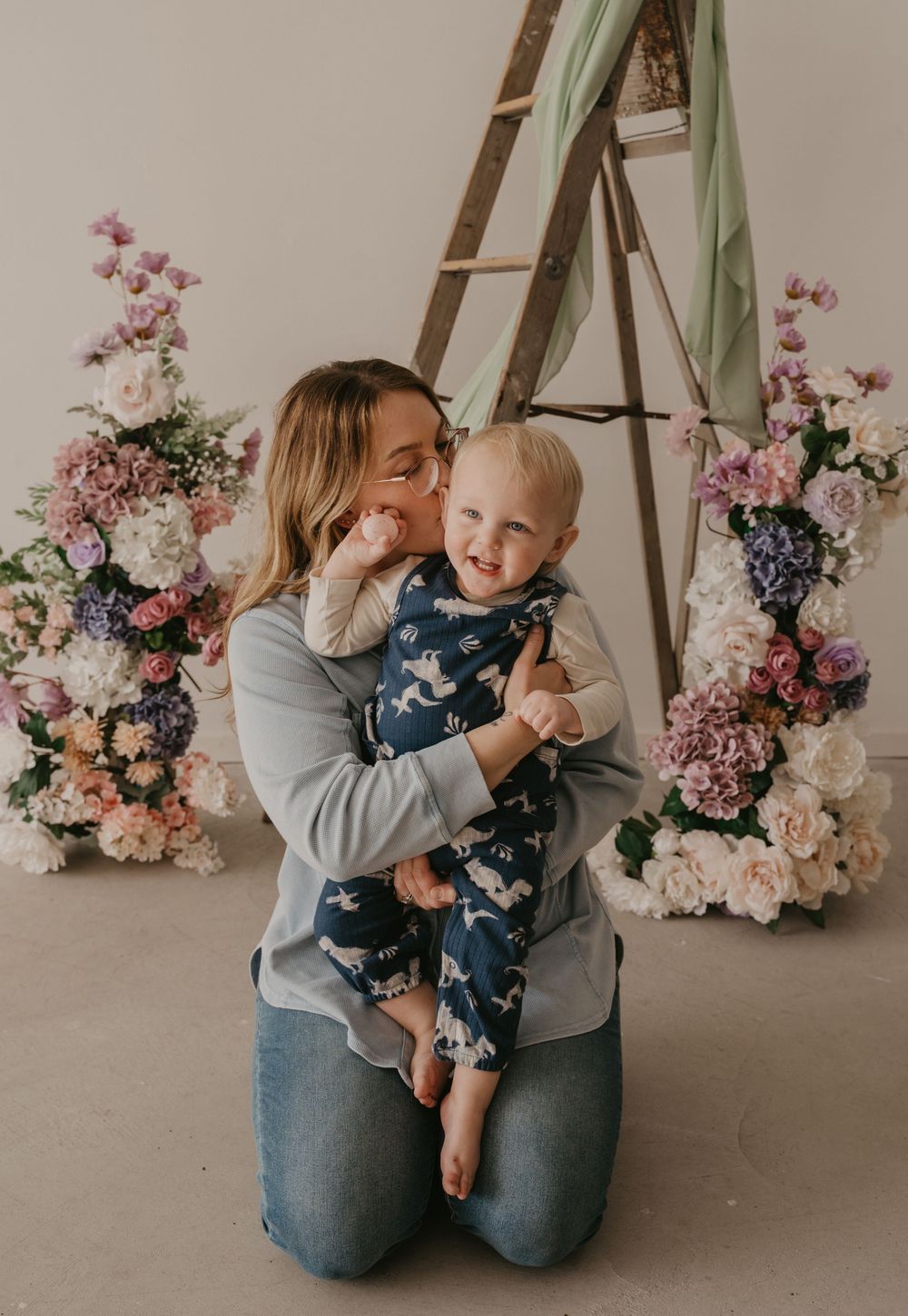 A mother kneeling and kissing her smiling baby dressed in blue pajamas, surrounded by floral arrangements in a studio.
