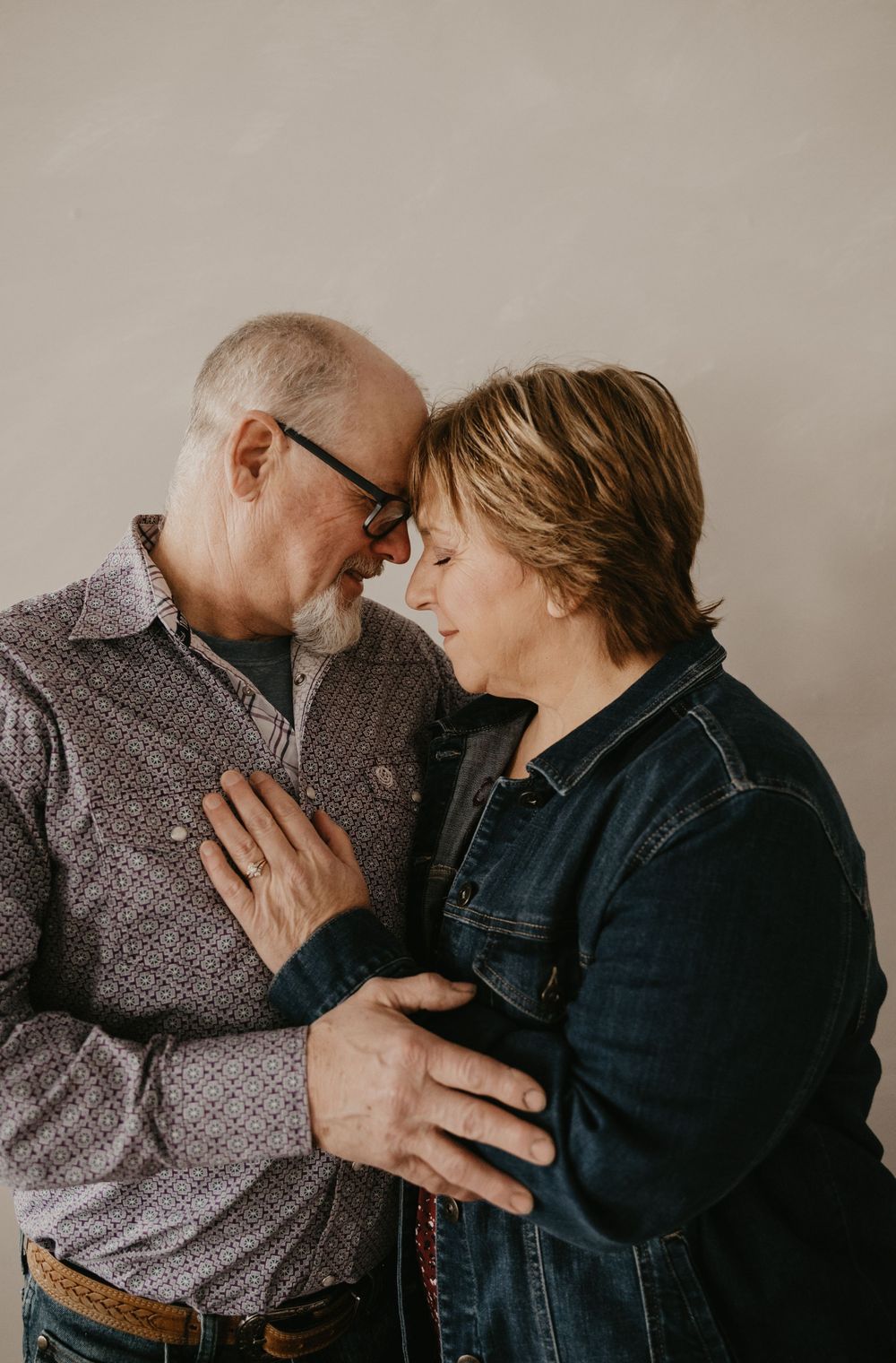 An older couple shares a tender forehead-to-forehead moment, smiling softly while embracing against a neutral background.