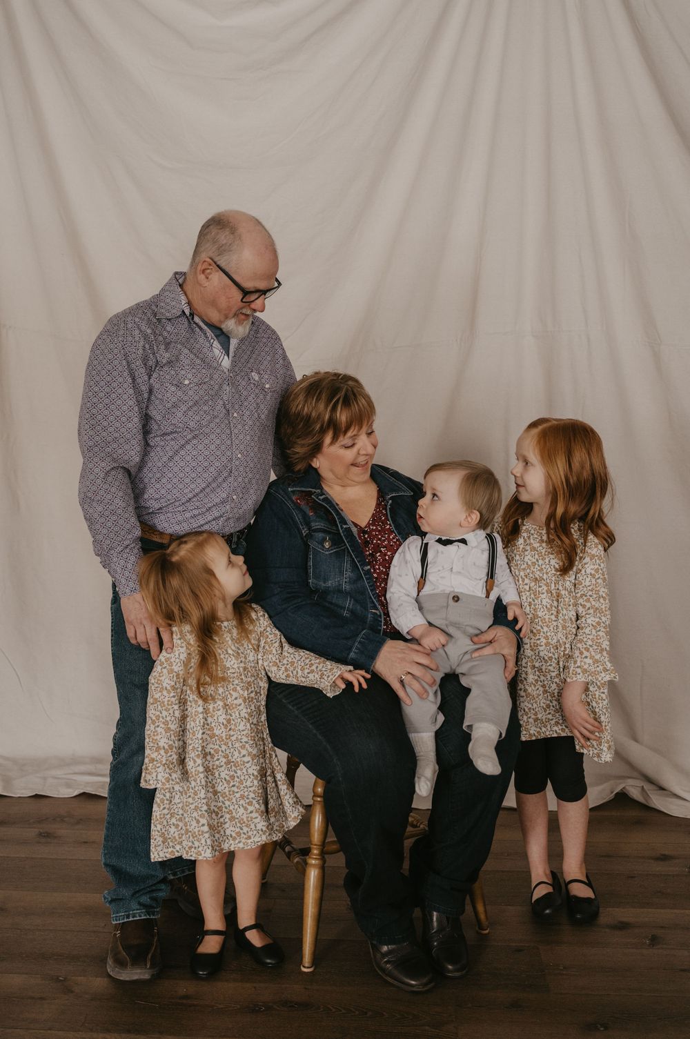 Grandparents posing with three young grandchildren, two girls in floral dresses and a baby boy, against a white backdrop.