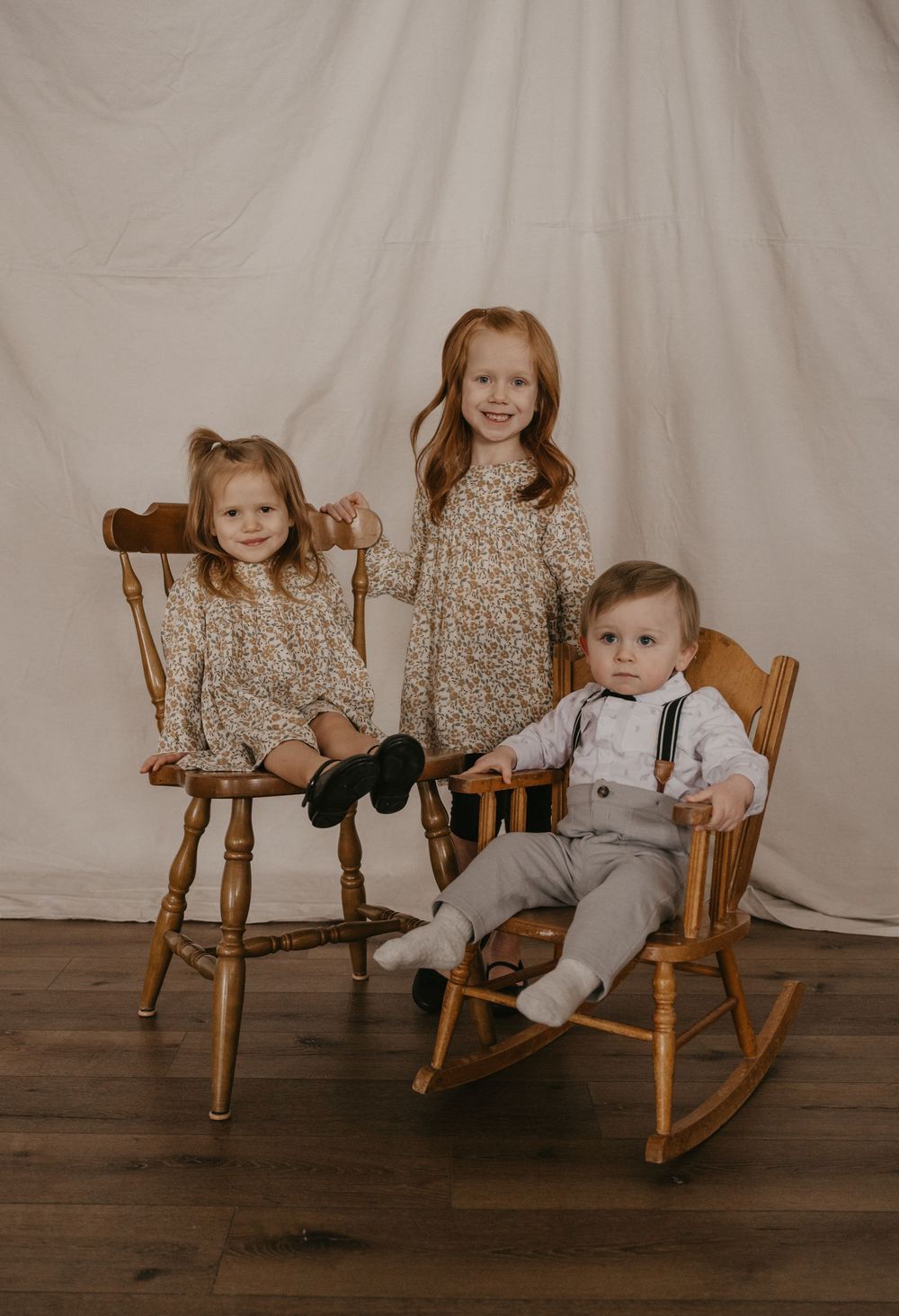 Three young children pose for a portrait, two girls in floral dresses and a baby boy in suspenders on wooden chairs.