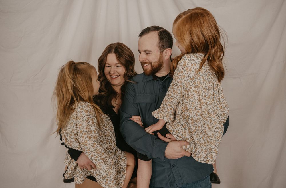 A happy family of four laughing together, two redhead girls in floral dresses with their smiling parents against a white backdrop.