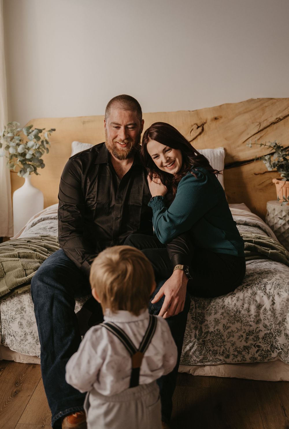 A smiling couple sits on a bed as their toddler in suspenders approaches them in a warmly lit bedroom.