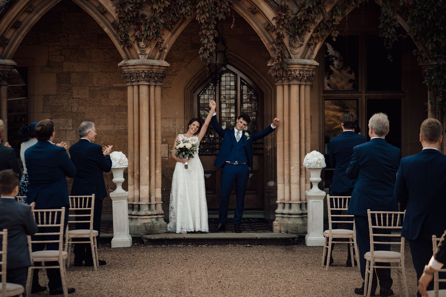 Wedding guests gather in an elegant stone archway corridor with wooden chiavari chairs at a formal event.