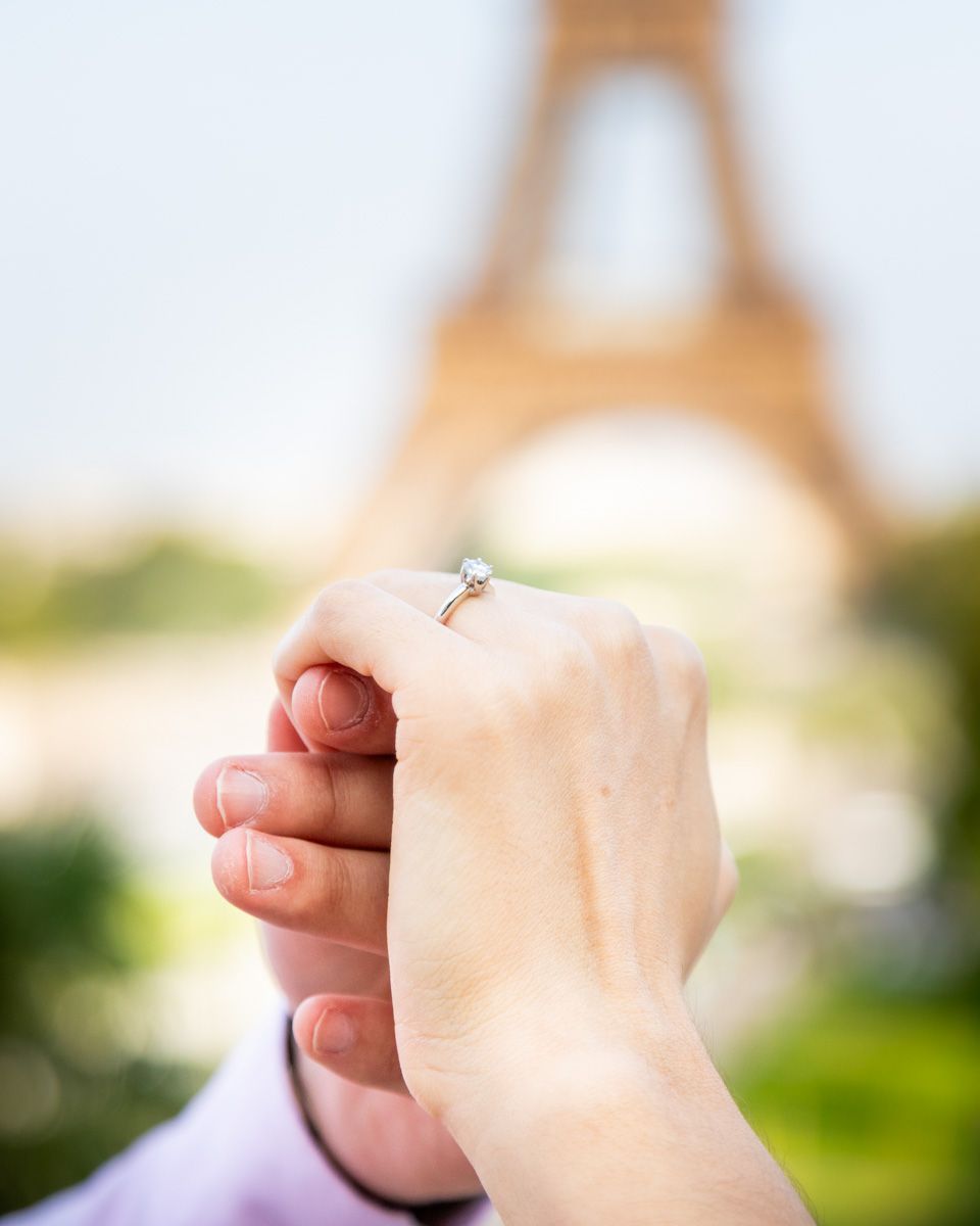A close-up photo of a hand with an engagement ring in front of the blurred Eiffel Tower in Paris.