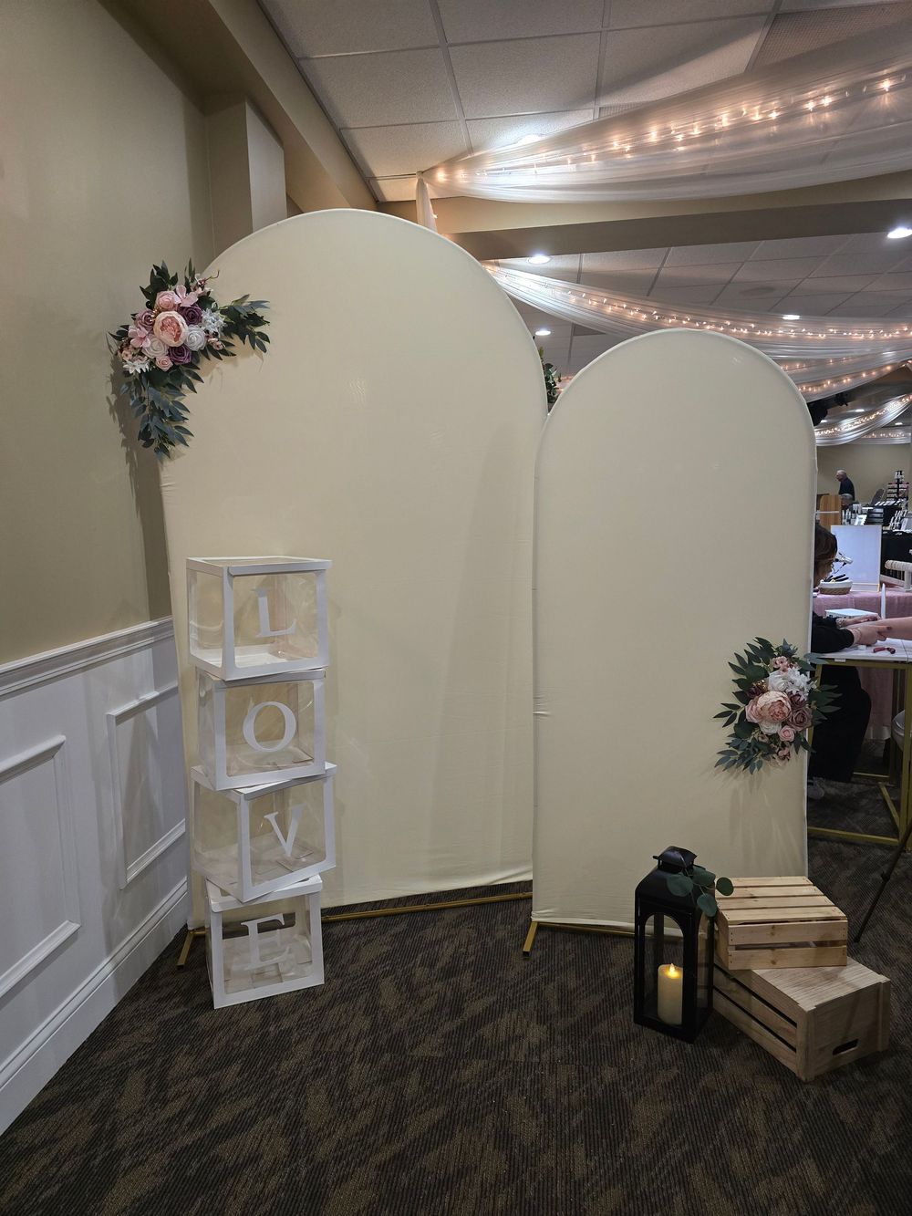 Elegant wedding backdrop with two ivory arch panels adorned with blush floral arrangements, LOVE letter boxes, lantern, and wooden crate.