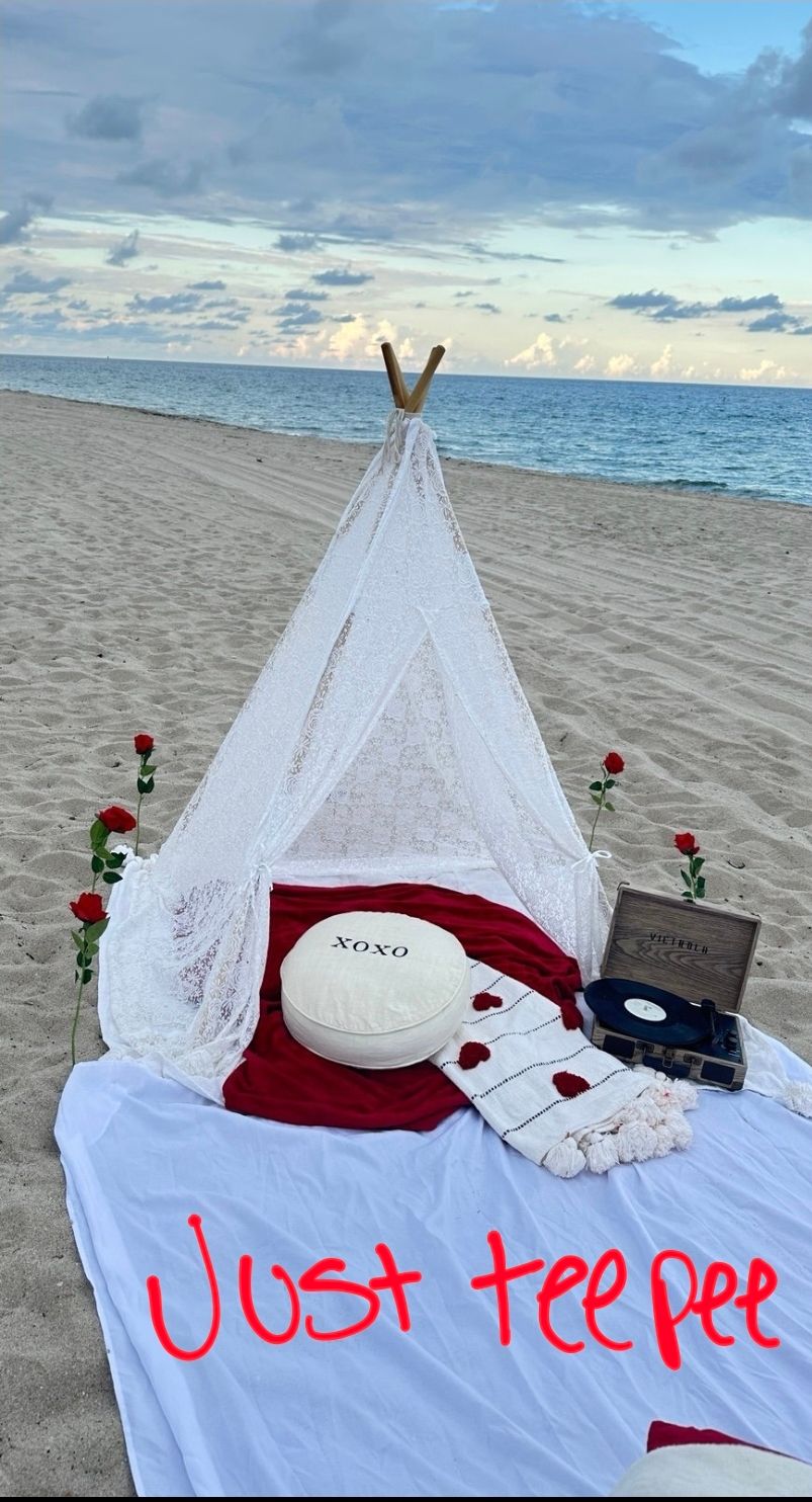 A romantic beach setup with a white teepee tent, red roses, and blankets on the sand at sunset.