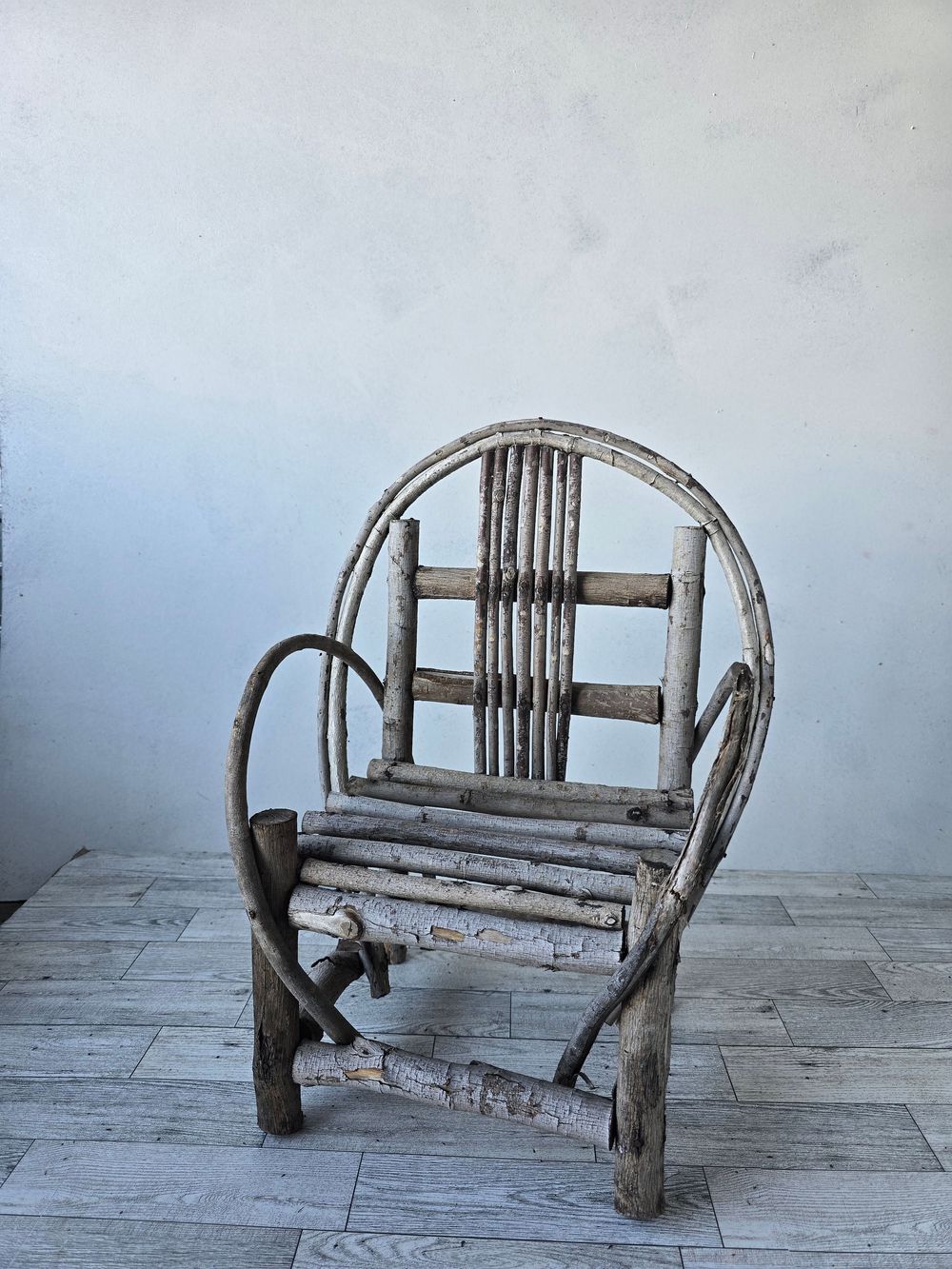 Rustic handmade twig chair with circular back and branch armrests, weathered gray wood, artisan folk art furniture.