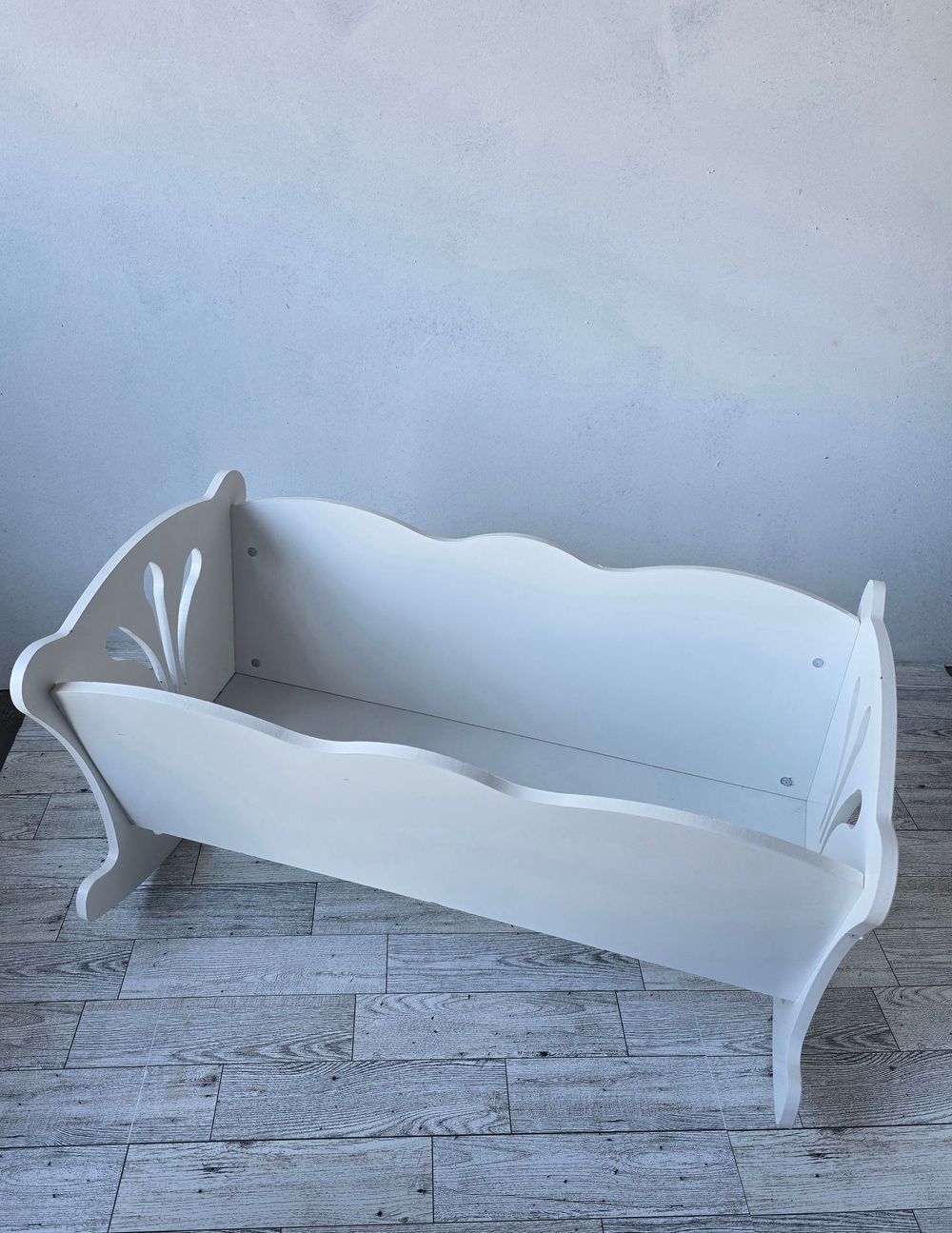 Vintage white wooden baby cradle with decorative cutout details and curved rockers on a light tile floor.