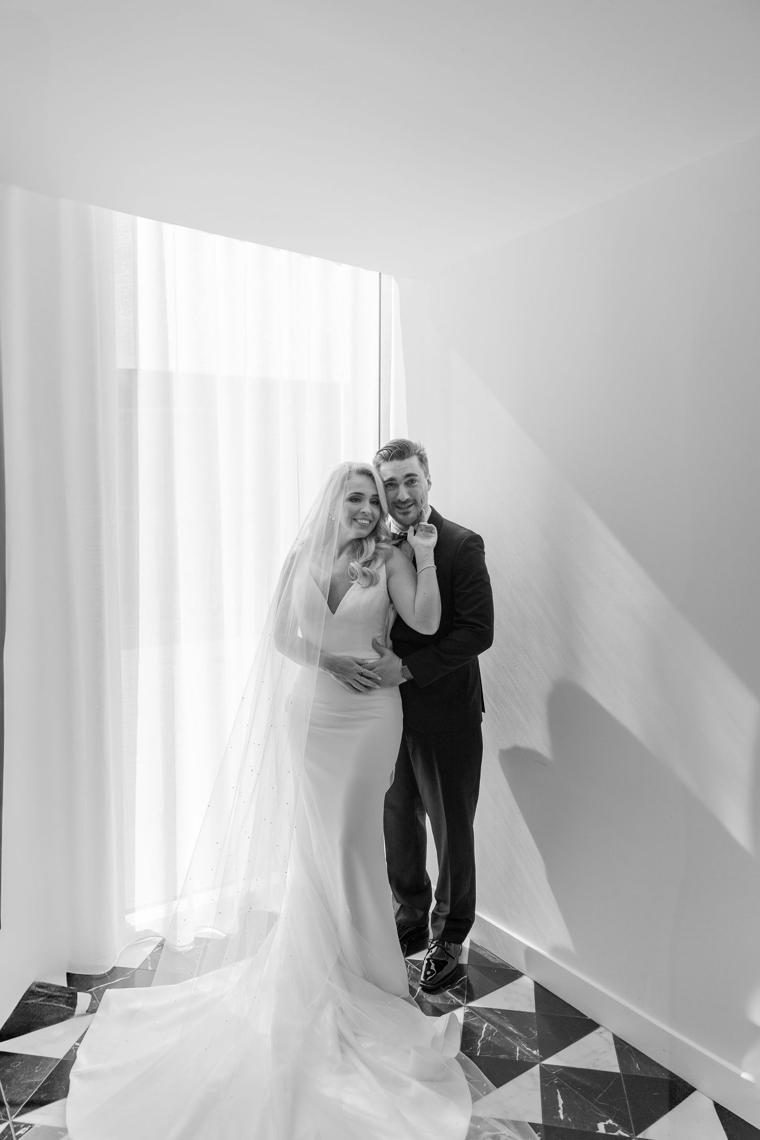 A romantic black and white wedding photo of a couple standing by a window with flowing curtains and geometric floor tiles. NASHVILLE WEDDING