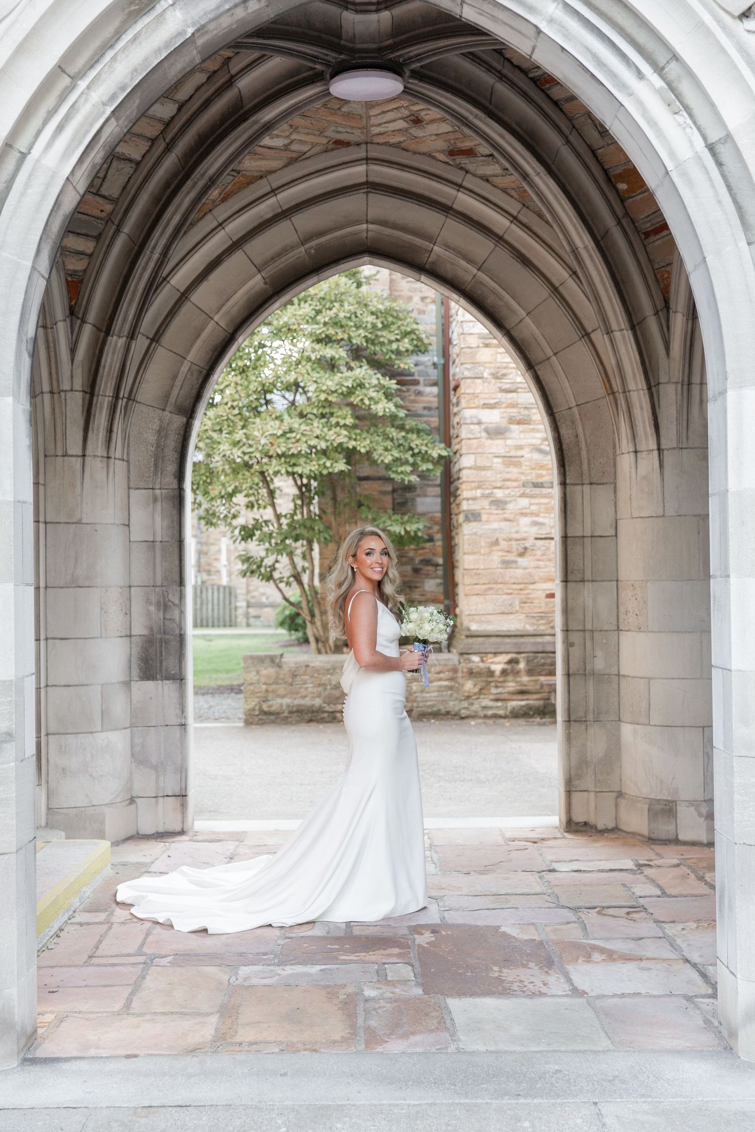 A bride in a fitted white gown poses under a gothic stone archway holding a bouquet. NASHVILLE WEDDING PHOTOGRAPHER