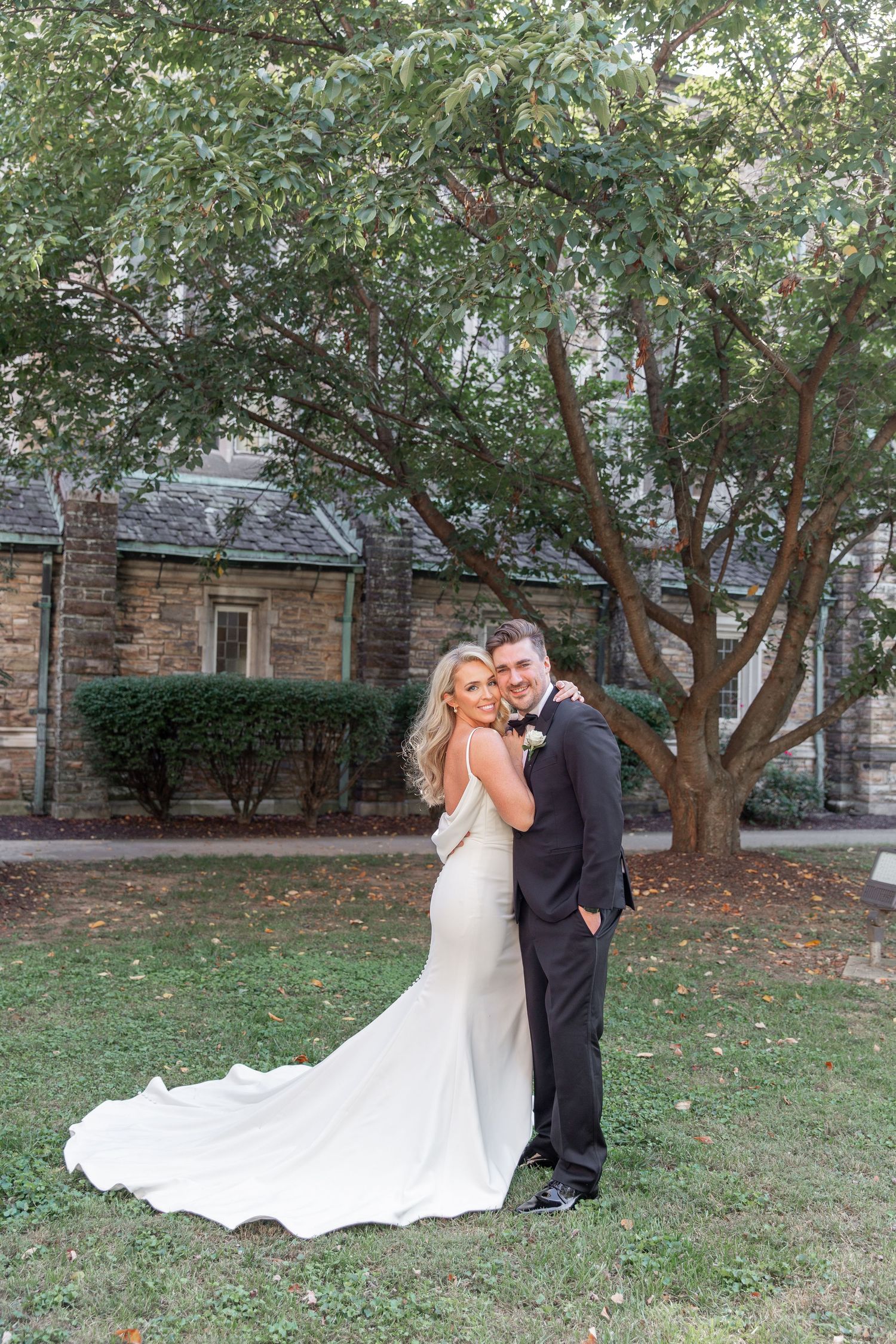 A couple embraces on their wedding day in front of a stone building with a large tree in the background. NASHVILLE WEDDING PHOTOGRAPHER