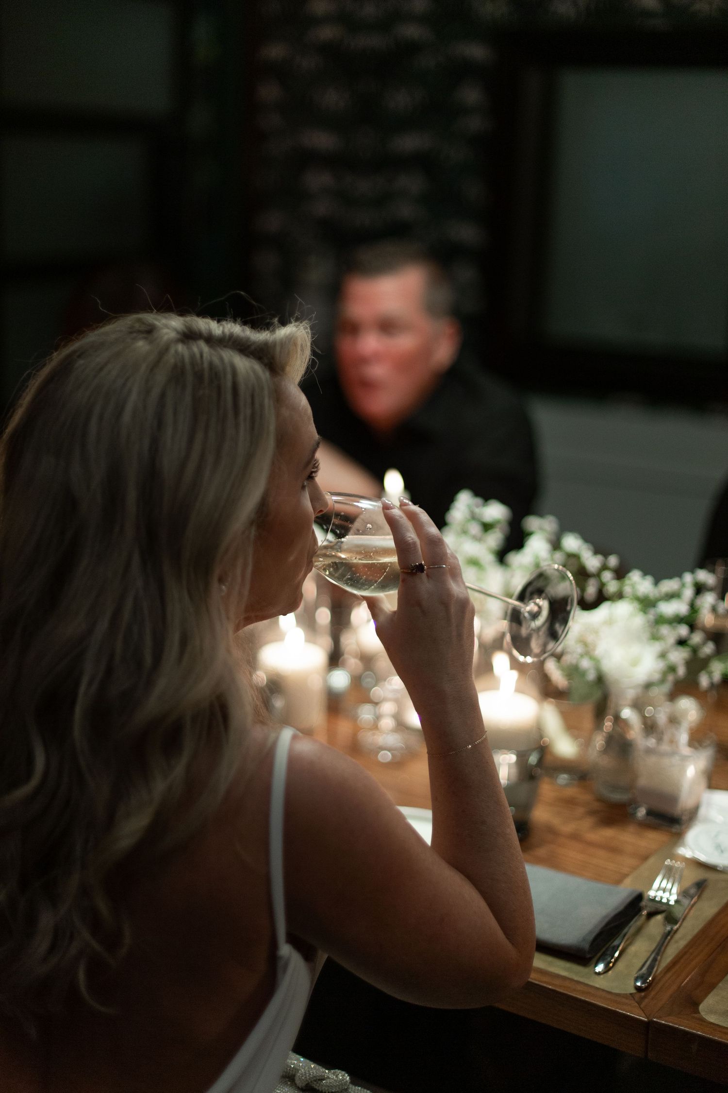 A person sits at a candlelit table with white floral arrangements and drinks from a wine glass in a dimly lit setting.NASHVILLE WEDDING PHOT