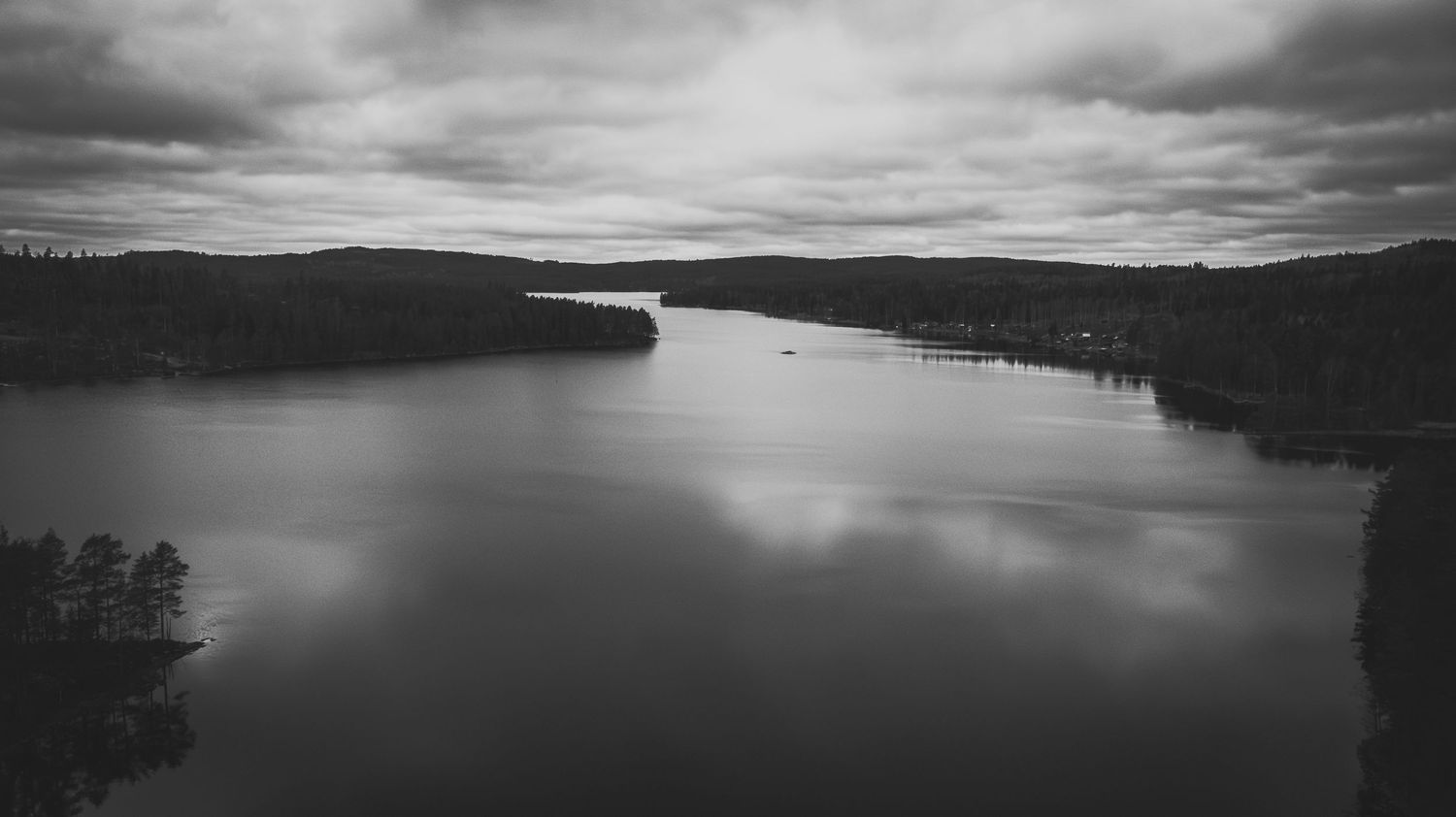 Svartvit bild av en stilla sjö med skog och molnig himmel