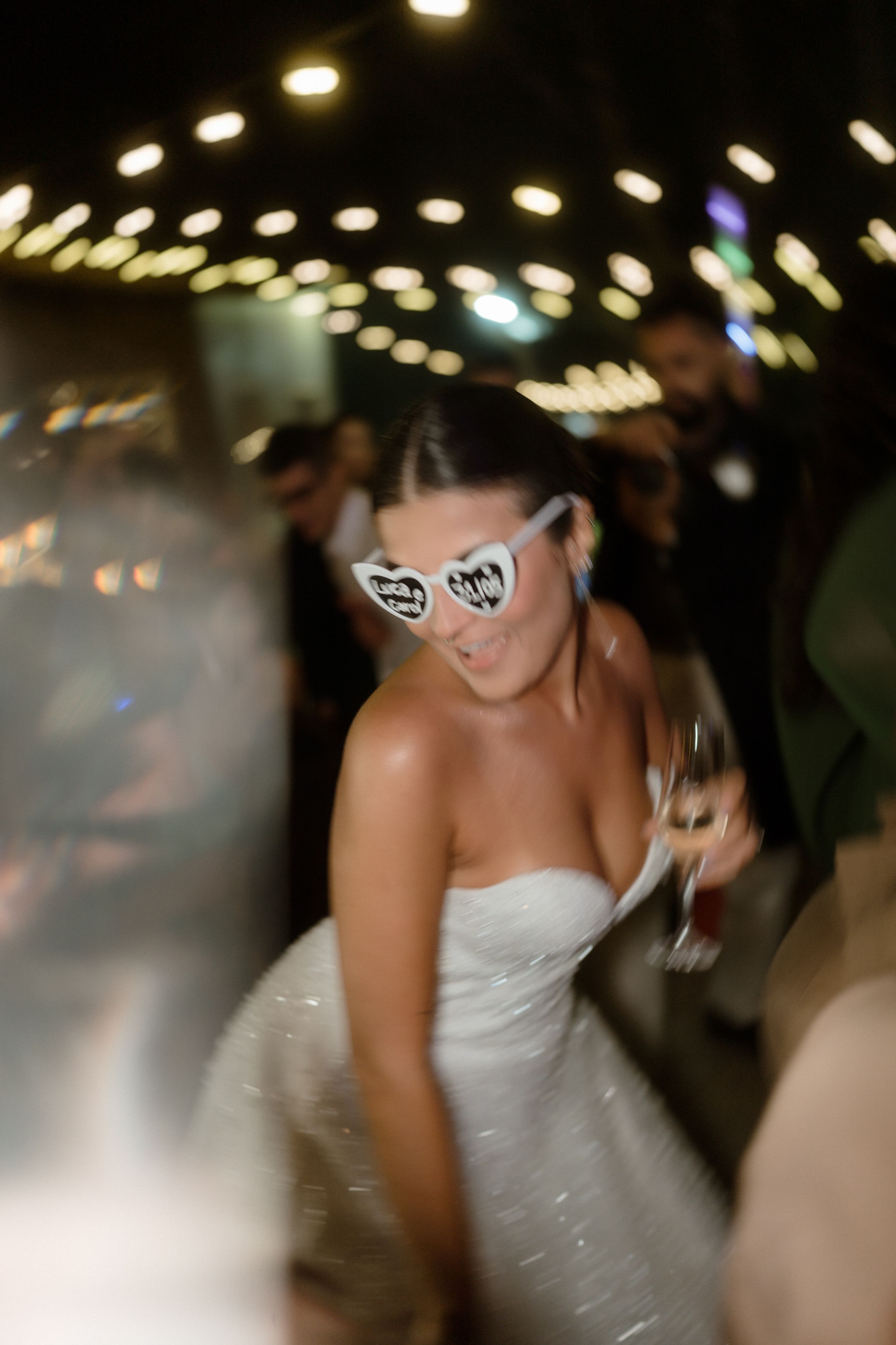Dancing figure in white dress and heart-shaped sunglasses under string lights at night celebration.