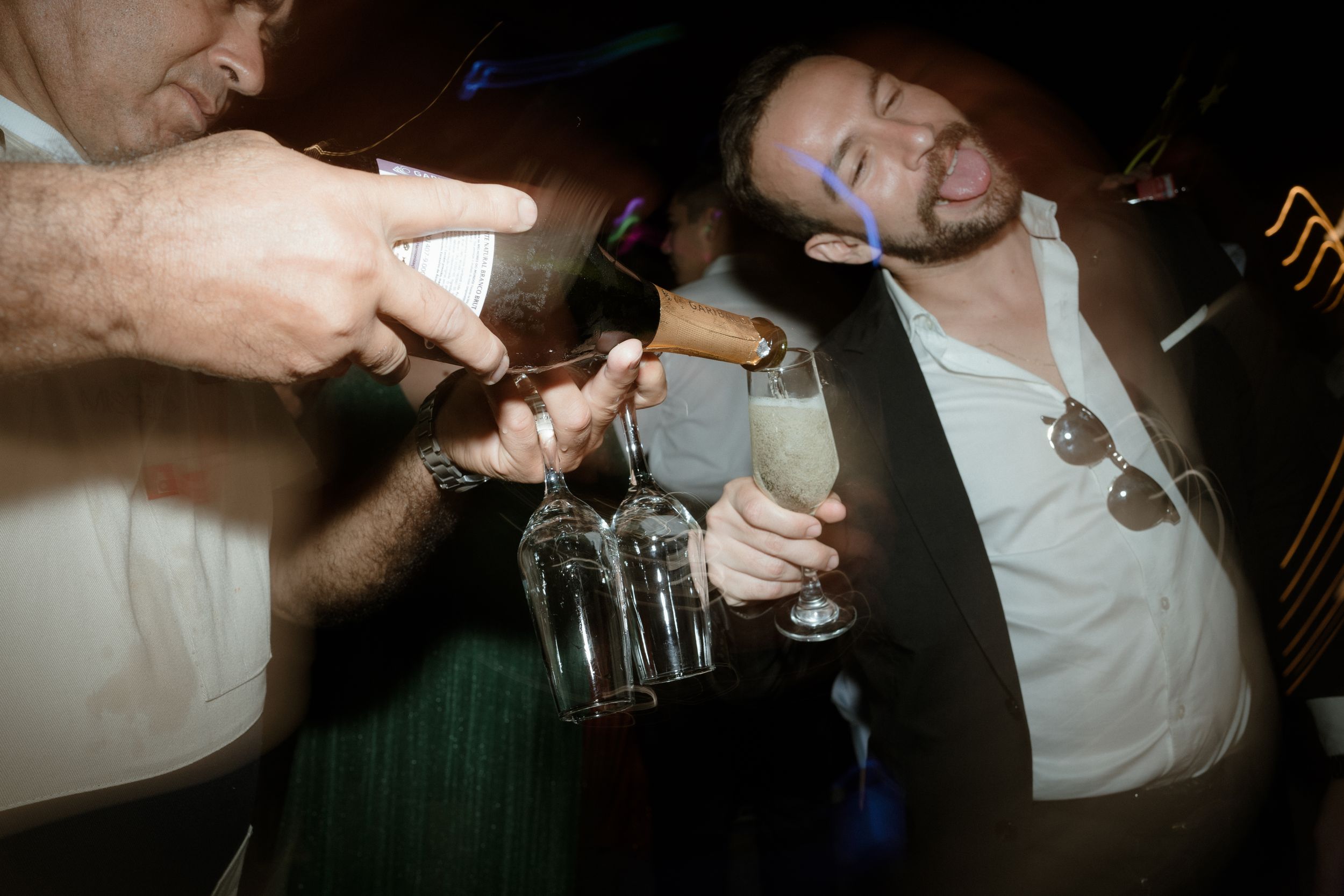 Party guests pouring drinks and toasting with bottles in dimly lit setting.