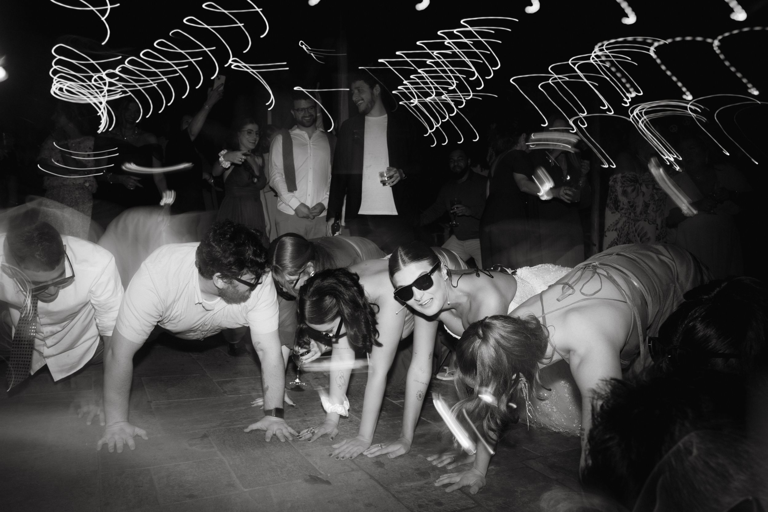 Black and white photo of wedding guests playing limbo game under string lights.