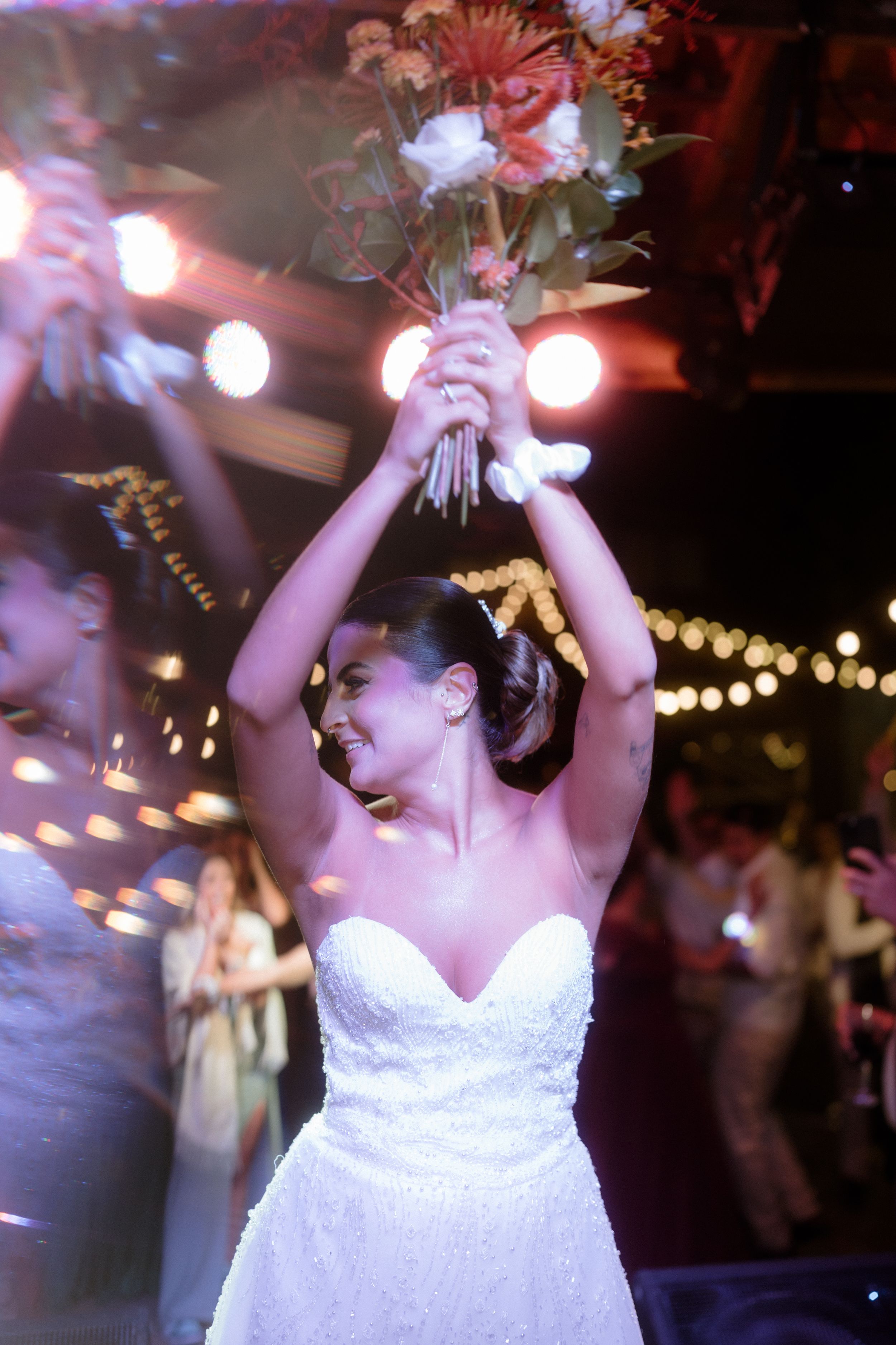 Bride in strapless white gown holds bouquet overhead during reception celebration.