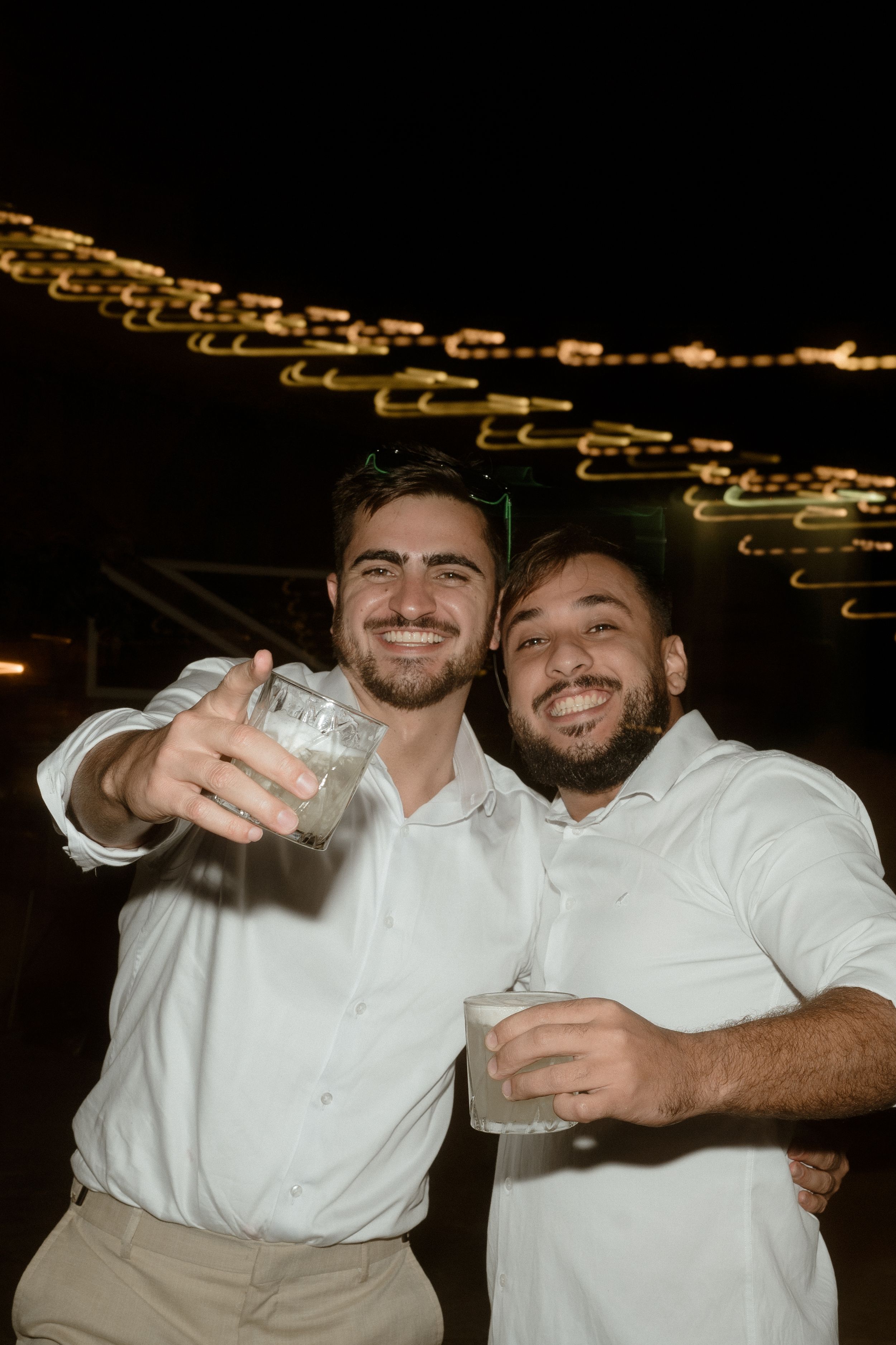 Two friends in white shirts toasting with drinks under string lights at night celebration.
