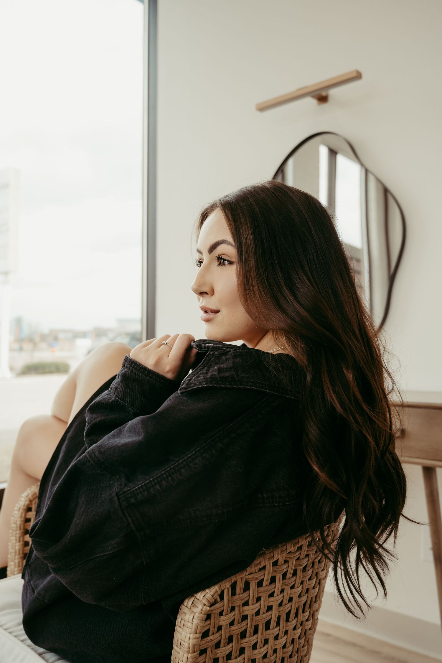Woman in black sweater sitting in woven chair by window in natural light.