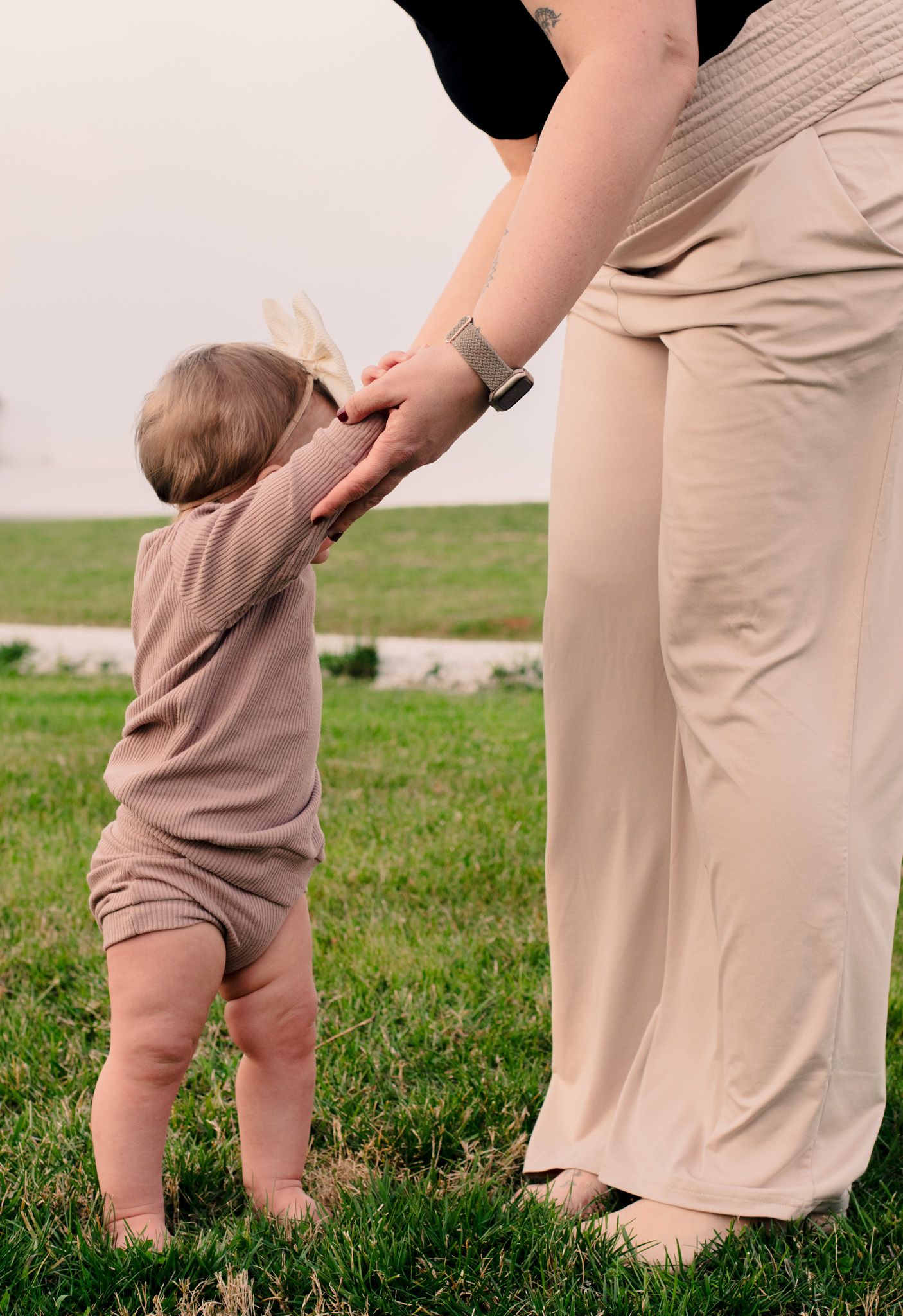 A small child in beige clothing reaches up to hold hands with an adult in a grassy field.