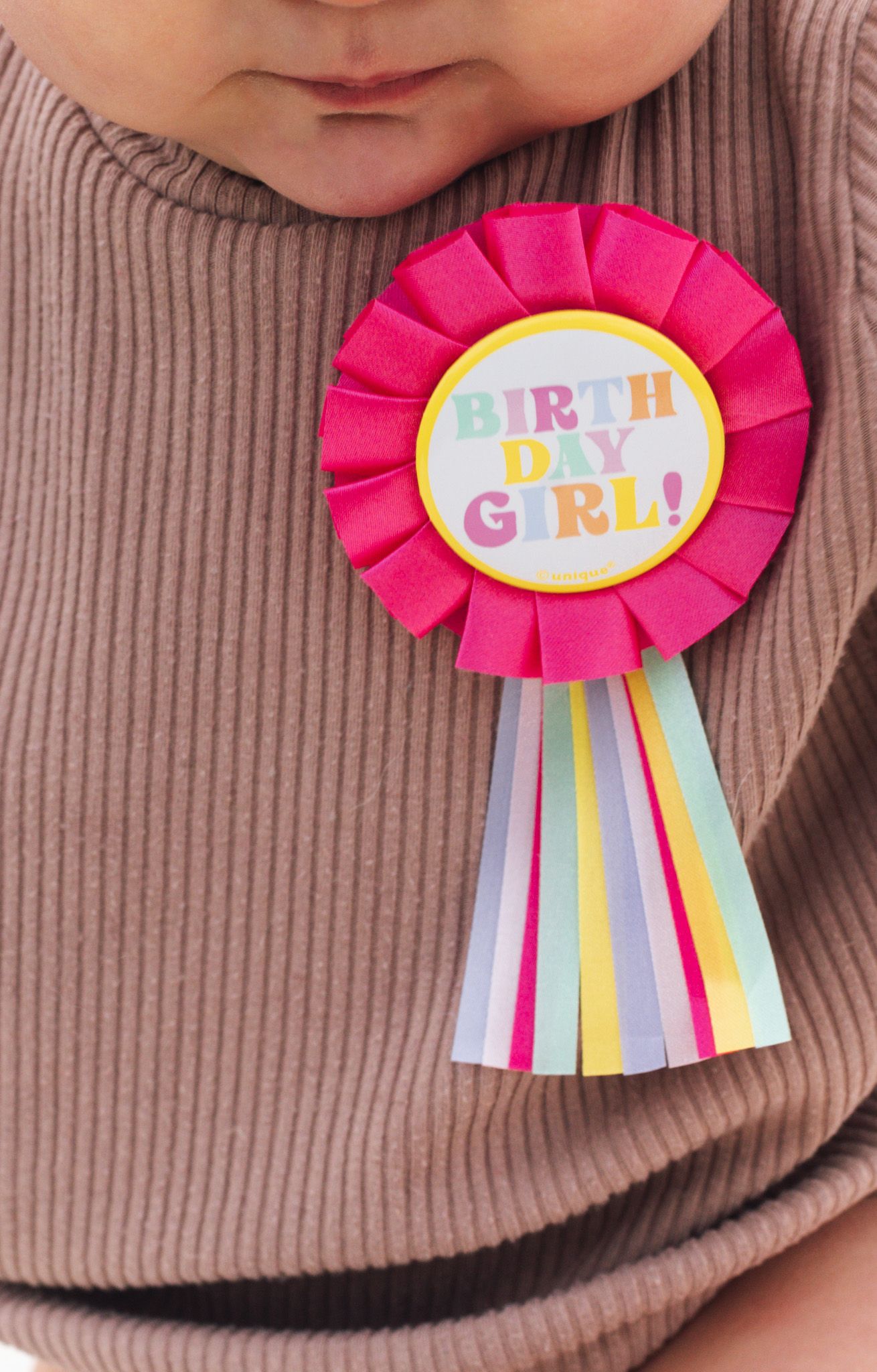 A pink birthday girl ribbon rosette with rainbow ribbons pinned to a brown sweater.