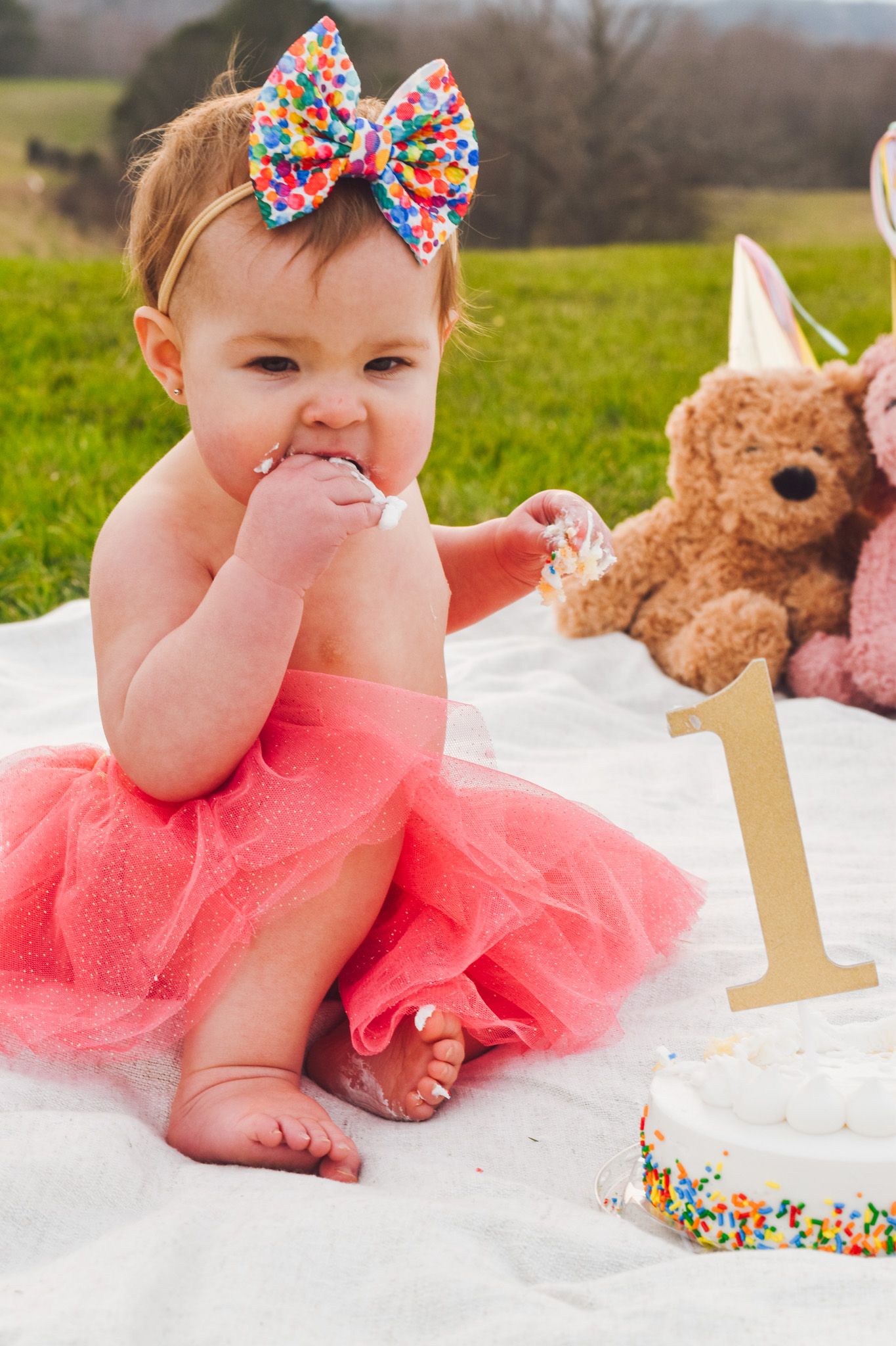 Baby in pink tutu and sparkly bow sits on blanket with teddy bear and number one decoration.