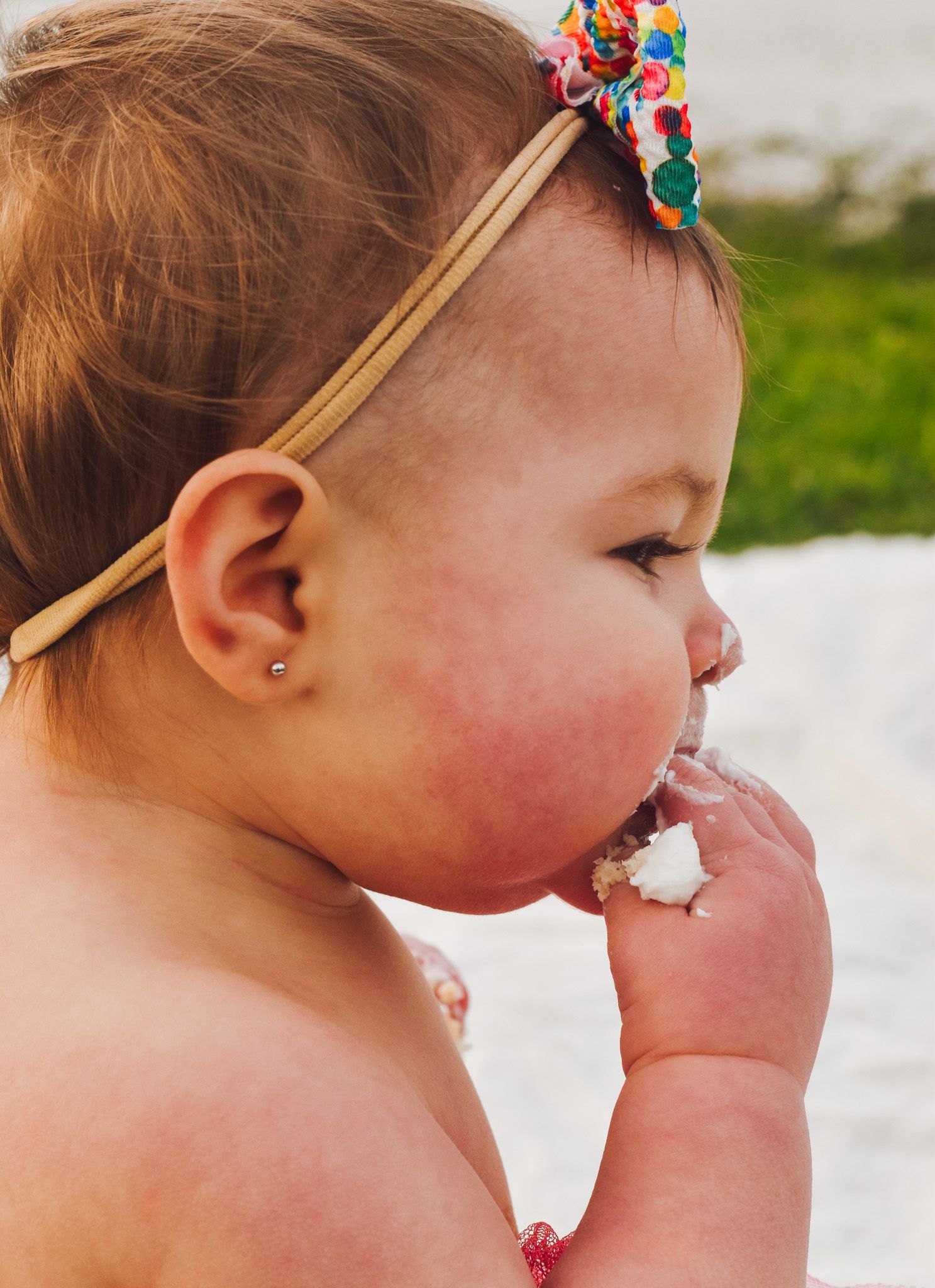 A baby wearing a colorful headband takes a bite of cake during a celebration.