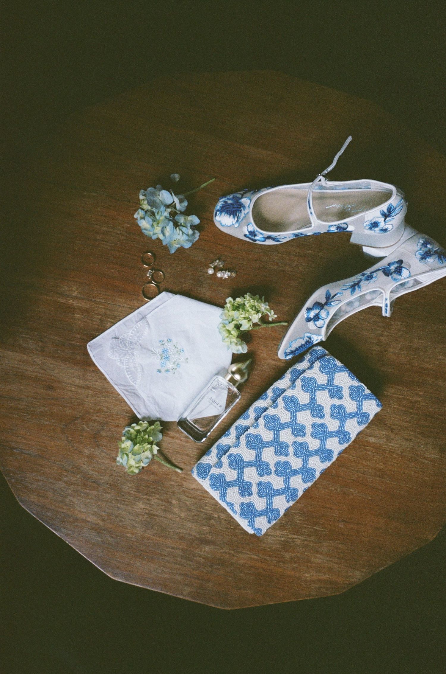 Wedding accessories including blue and white floral shoes, handkerchief, and small flowers on wooden surface.